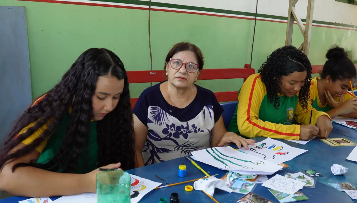 Mulher de óculos vermelhos e blusa estampada, sentada à mesa entre estudantes que pintam desenhos, observa atentamente o trabalho ao seu lado.