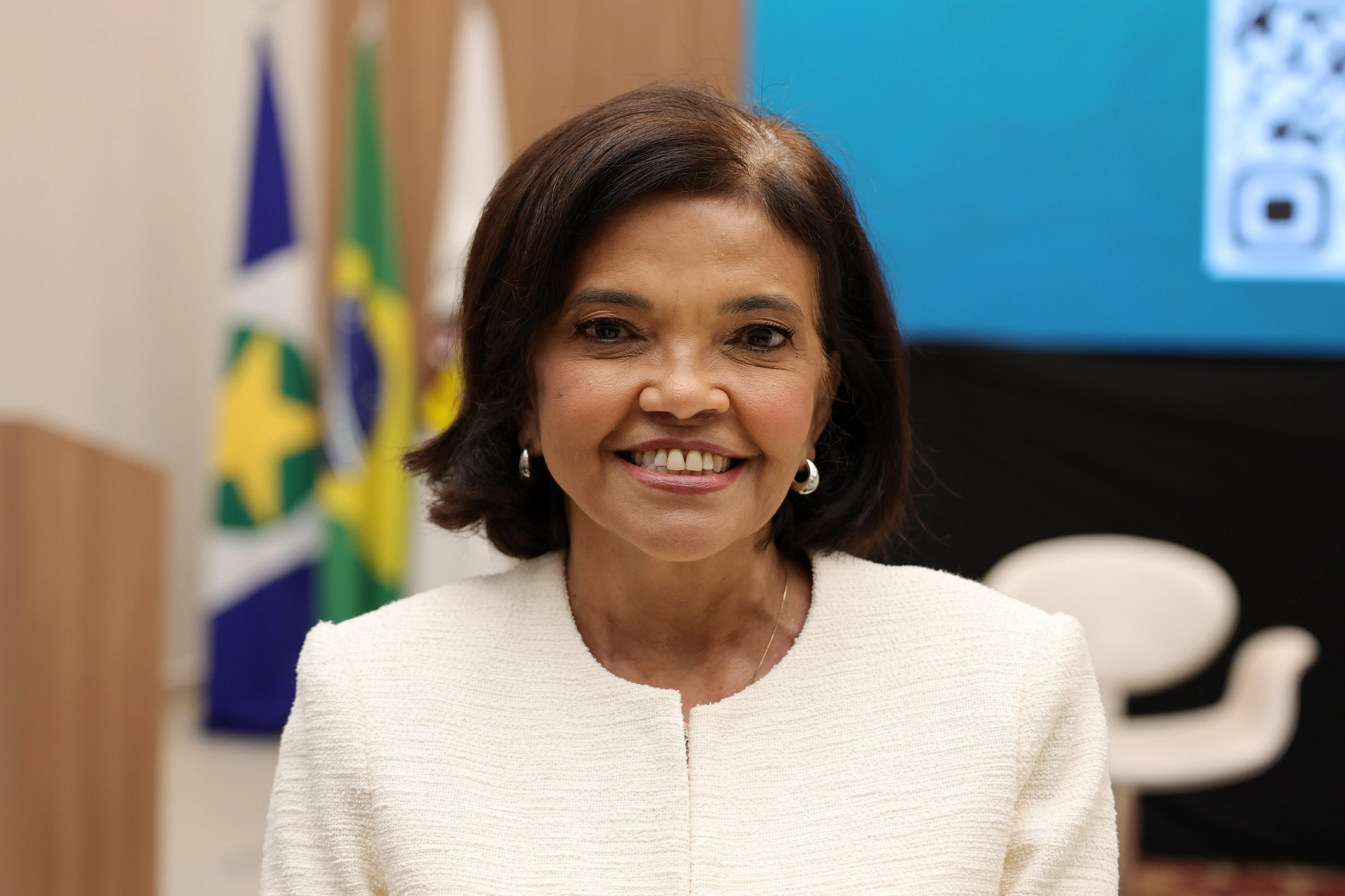 Plano médio de mulher negra e cabelos escuros curtos, sorrindo de frente. Usa casaco bege e brincos de argola pequenos. Ao fundo desfocado, veem-se as bandeiras de Mato Grosso e do Brasil em mastros.