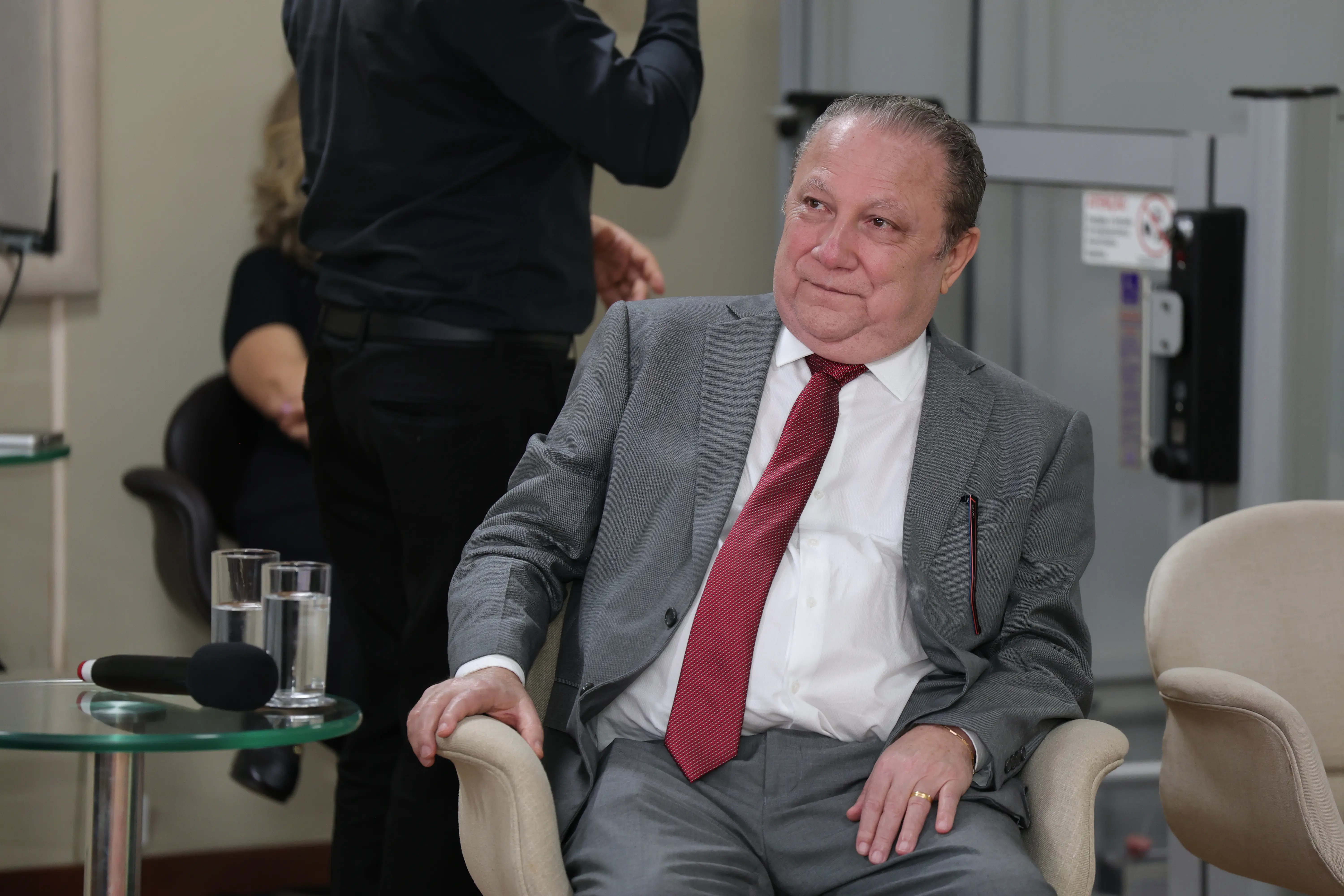 Um homem de terno cinza e gravata vermelha está sentado em uma poltrona, olhando para o lado. Ao fundo, uma mesa com copos de água e uma pessoa desfocada.