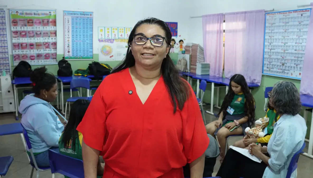 Mulher de cabelos escuros, óculos e blusa vermelha, posa em sala de aula decorada com cartazes pedagógicos, enquanto alunas e adultos conversam ao fundo.