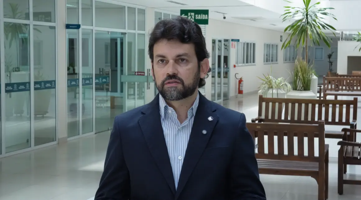 Homem de cabelos escuros, barba e terno azul-marinho sobre camisa listrada, é fotografado em corredor amplo com plantas e bancos de madeira.