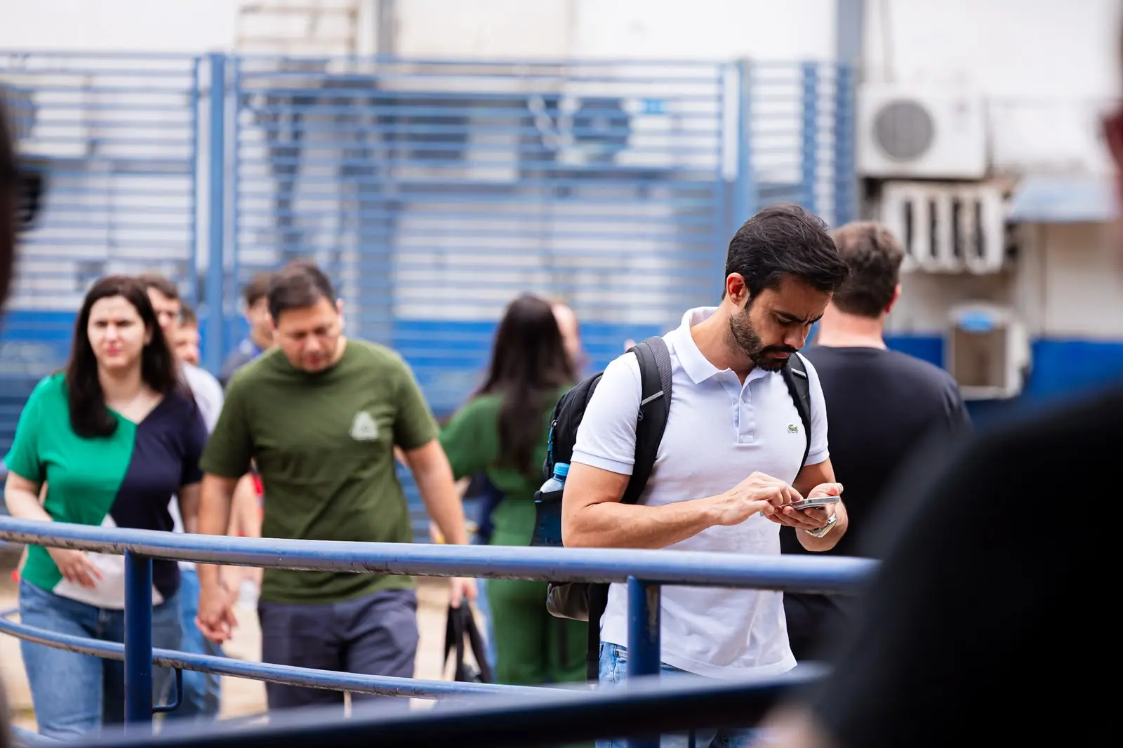 Homem de pele clara, cabelos escuros e barba curta veste camisa polo branca e carrega uma mochila preta. Ele está em primeiro plano, caminhando e mexendo no celular. Ao fundo, um portão azul e outras pessoas se movimentam em um ambiente externo.