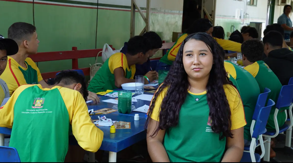 Jovem de cabelos longos e cacheados, veste camiseta verde e amarela, observa a câmera em pátio escolar onde outros estudantes desenham à mesa.