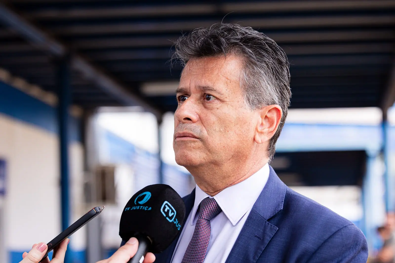 Homem de meia-idade, cabelos escuros grisalhos e terno azul com gravata estampada, olha para o lado com expressão séria. Ele está sendo entrevistado com um microfone preto da "TV JUS" apontado em sua direção.