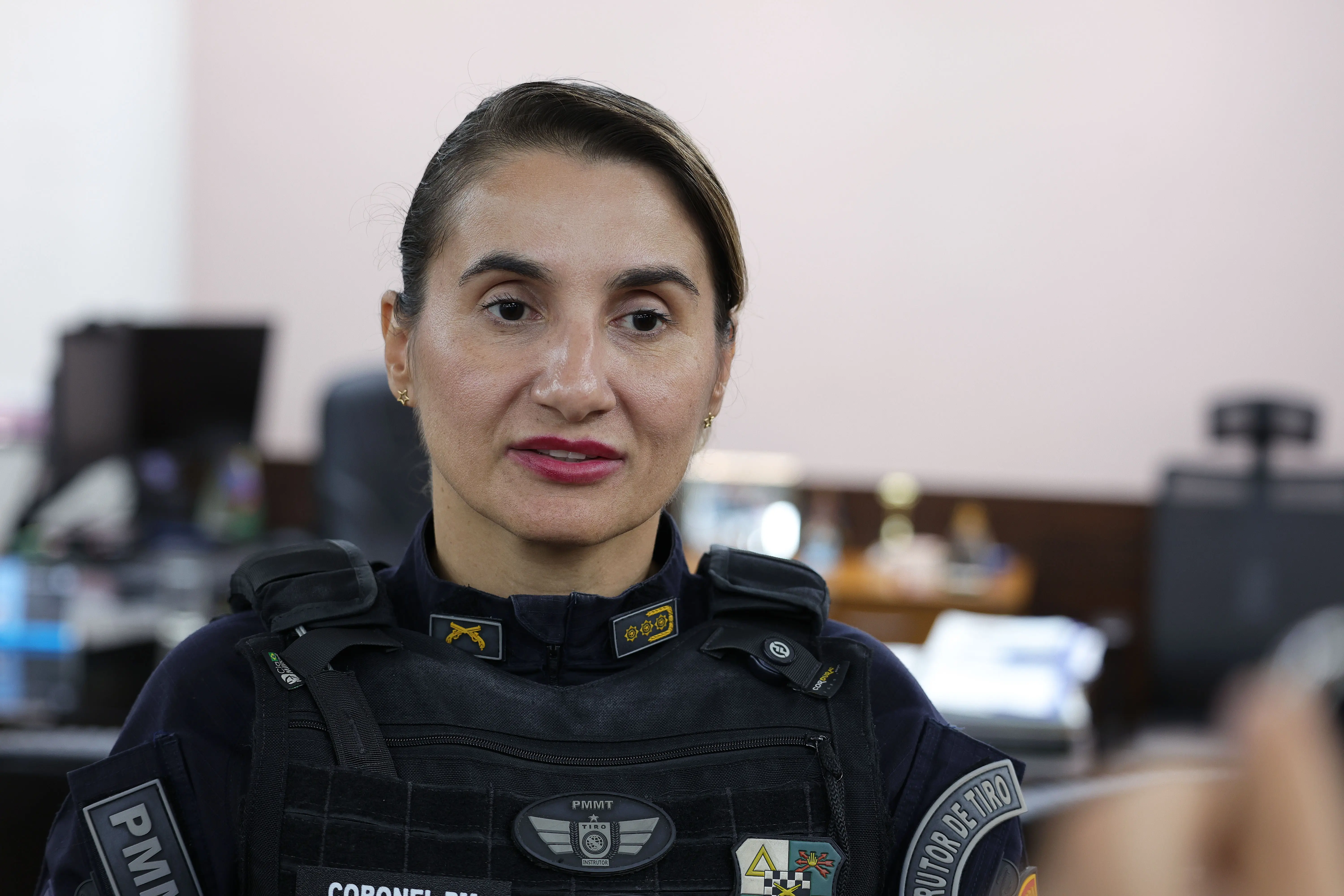 Subchefe de Estado-Maior Geral da Polícia Militar, coronel PM Grasielle Paes usa uniforme da PM, incluindo um colete à prova de balas preto com o emblema da "PMMT", tem cabelo escuro e  preso