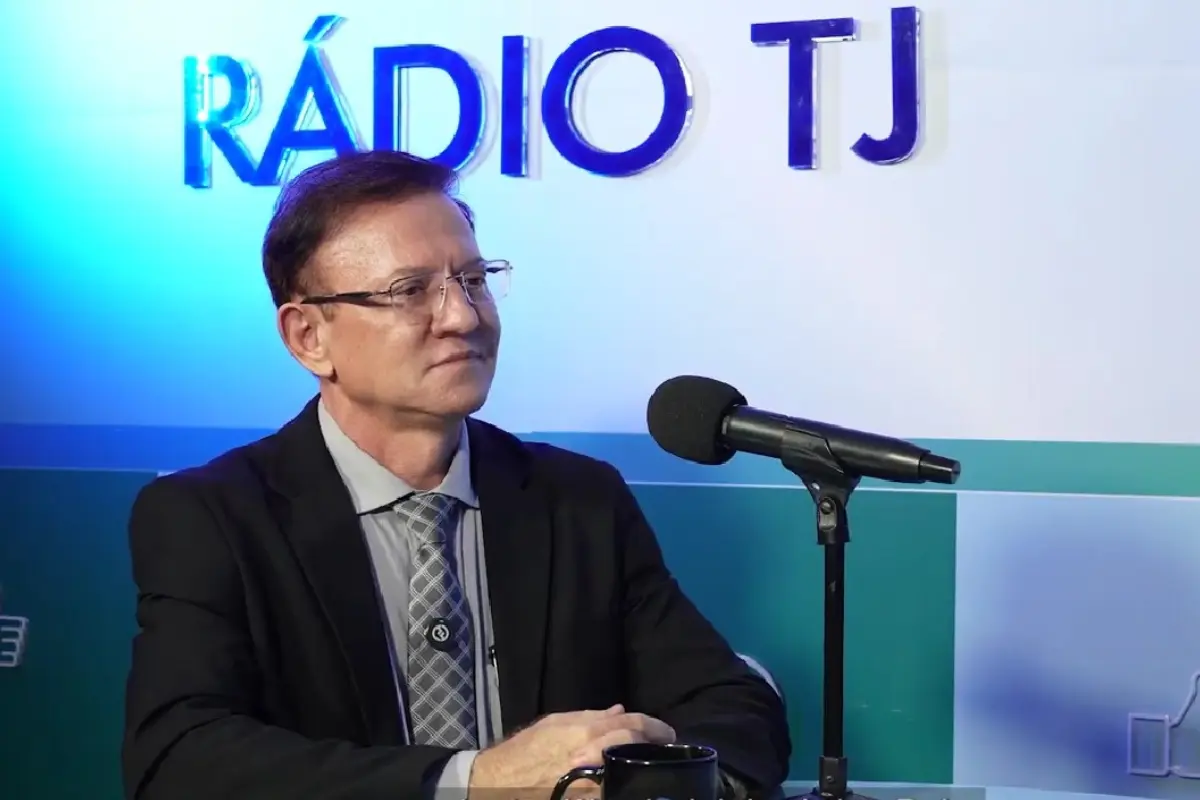 Homem de óculos, cabelos curtos e escuros, vestindo terno preto, camisa cinza claro e gravata estampada, está sentado diante de um microfone. Ao fundo, uma parede com o logotipo iluminado "RÁDIO TJ"