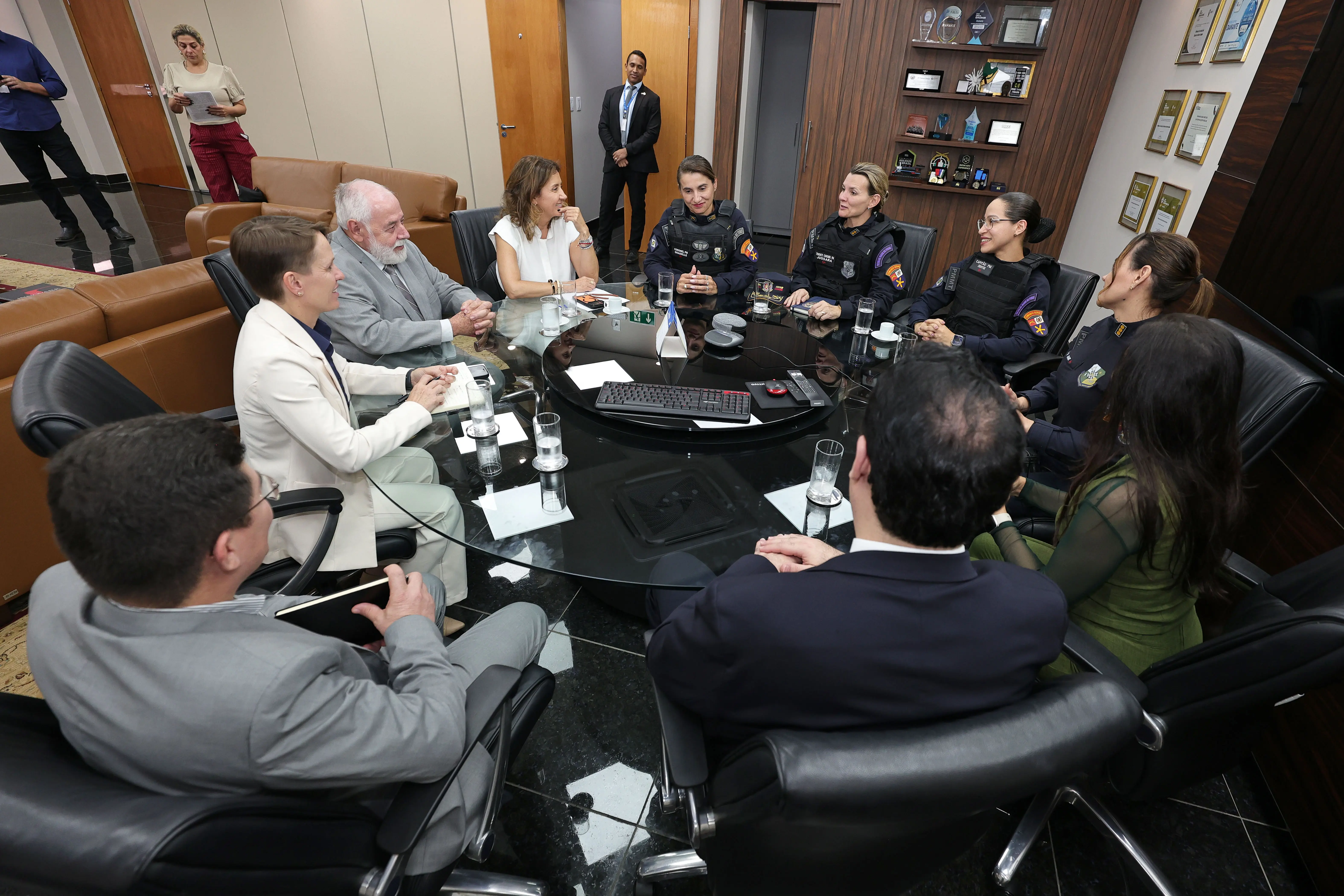 Em um escritório, um grupo de pessoas, incluindo policiais fardados e civis de terno e blazer, está sentado ao redor de uma mesa redonda de vidro em uma reunião.