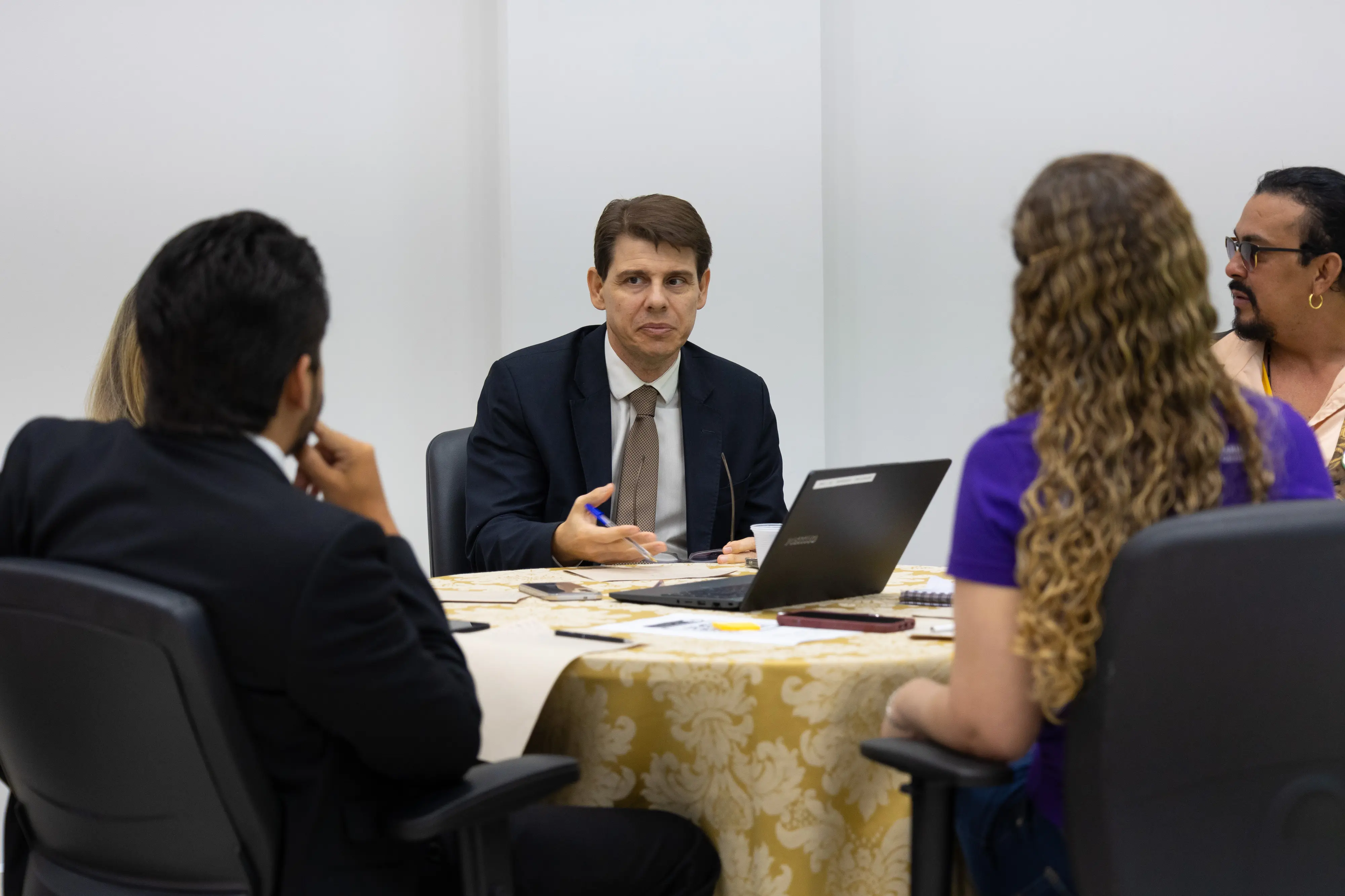 Homem de terno conversa com participantes sentados em mesa redonda durante atividade institucional.