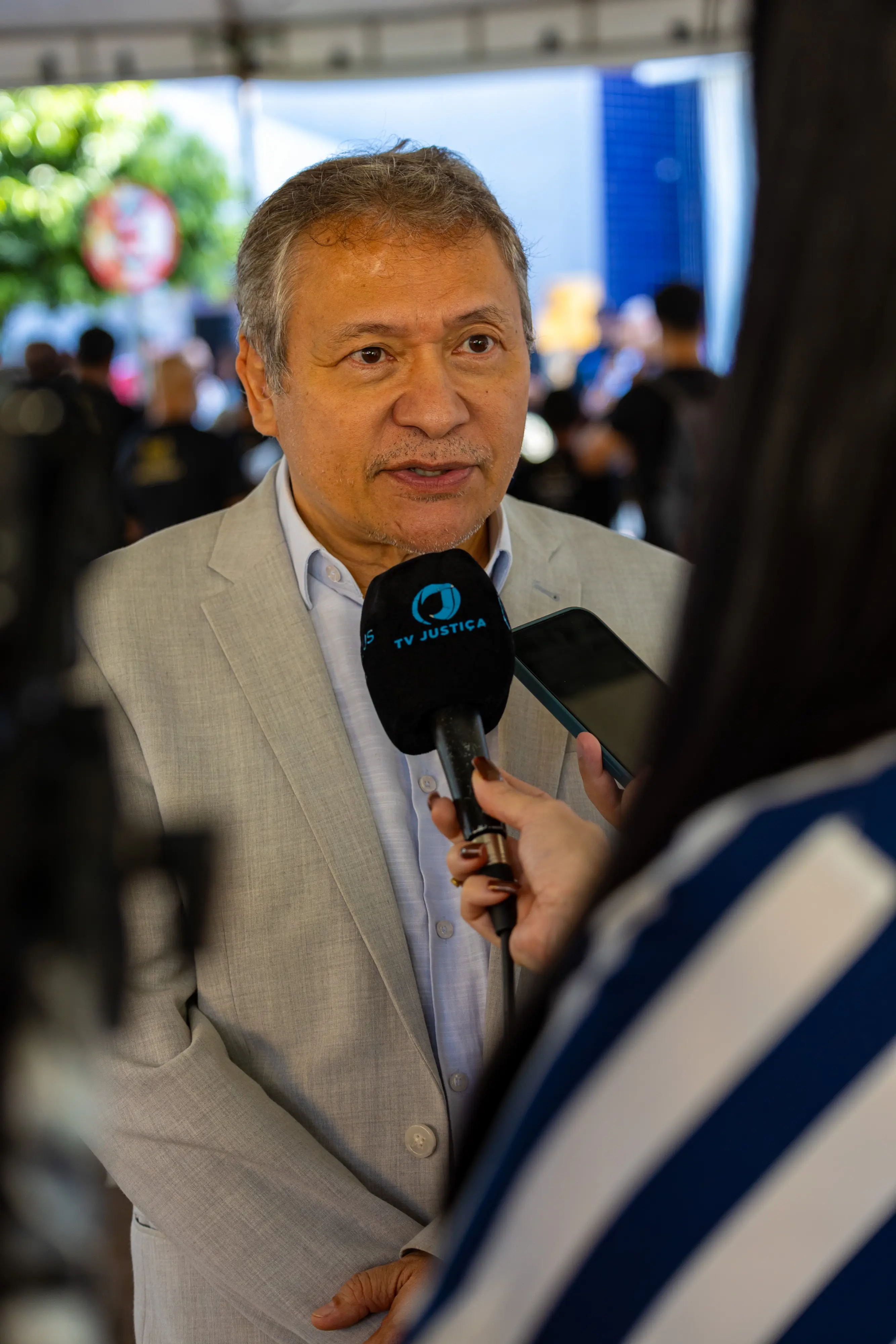 Homem de cabelos grisalhos, veste terno cinza e concede entrevista ao canal TV Justiça. Ele olha para a repórter com expressão serena e profissional.