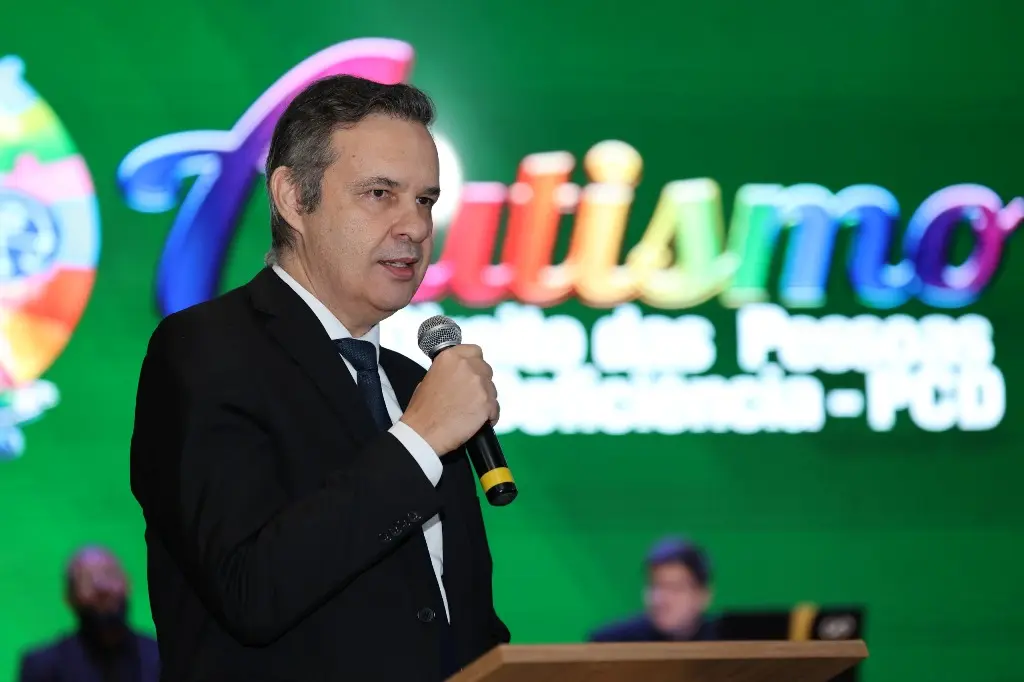Foto horizontal colorida que mostra o juiz auxiliar da Presidência do TJMT, Agamenon Alcântara, falando ao microfone, no púlpito. ele é um homem branco, alto, de cabelos grisalhos, usando camisa branca, terno e gravata pretos. Atrás dele há um enorme telão com a logomarca do evento TJMT Inclusivo.
