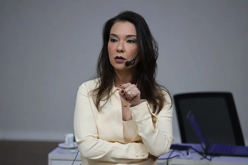 Foto horizontal em plano médio que mostra a juíza Hanae Yamamura proferindo palestra. Ela é uma mulher branca, de traços orientais, cabelos castanhos, lisos e soltos, usando blusa e blazer cor creme.