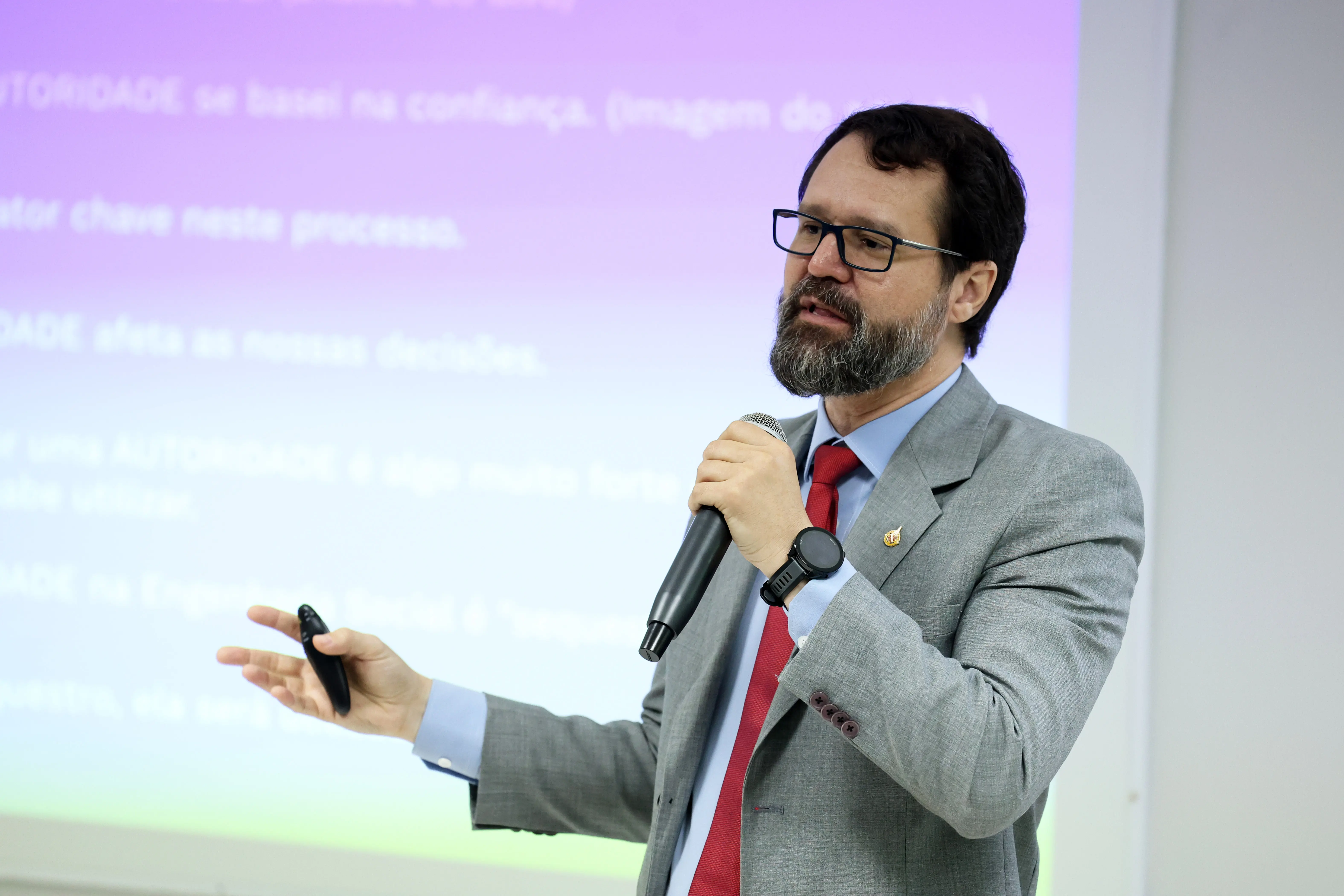 Professor do curso, André Valdevino. Ele tem pele clara, com barba e bigode grisalhos e bem aparados, usando óculos de armação preta. Ele veste um paletó cinza claro, camisa azul clara e uma gravata vermelha.