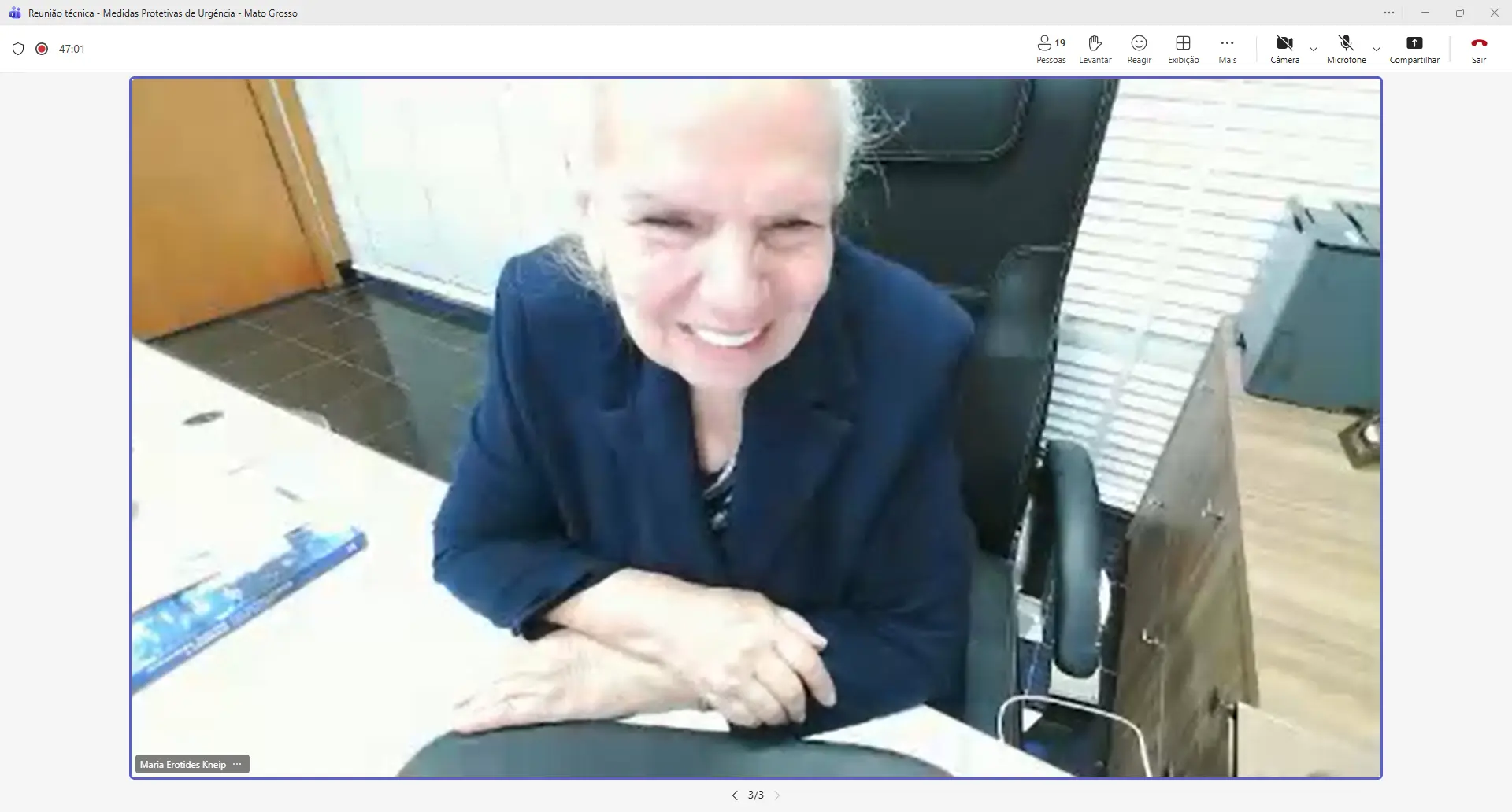 Mulher de cabelos brancos compridos, veste blazer azul marinho, sorri com as mãos entrelaçadas sobre a mesa. Ao fundo, cadeira de escritório e persianas brancas.