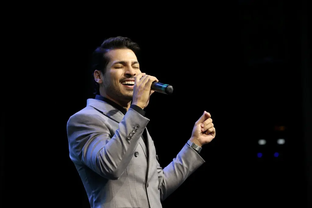 Foto horizontal colorida em plano fechado que mostra o cantor Tiago Roseno cantando de olhos fechados e sorrindo. ele é um homem jovem, branco, de cabelos escuros, usando terno cinza. 
