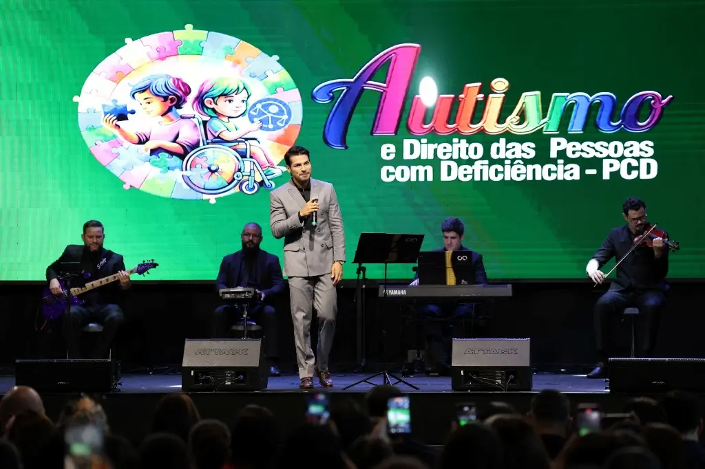 Foto horizontal colorida em plano aberto que mostra o cantor Tiago Roseno e músicos da Cia Sinfônica se apresentando no palco do evento TJMT Inclusivo. No telão aparece a logomarca do evento e, na parte inferior da foto, parte do público.