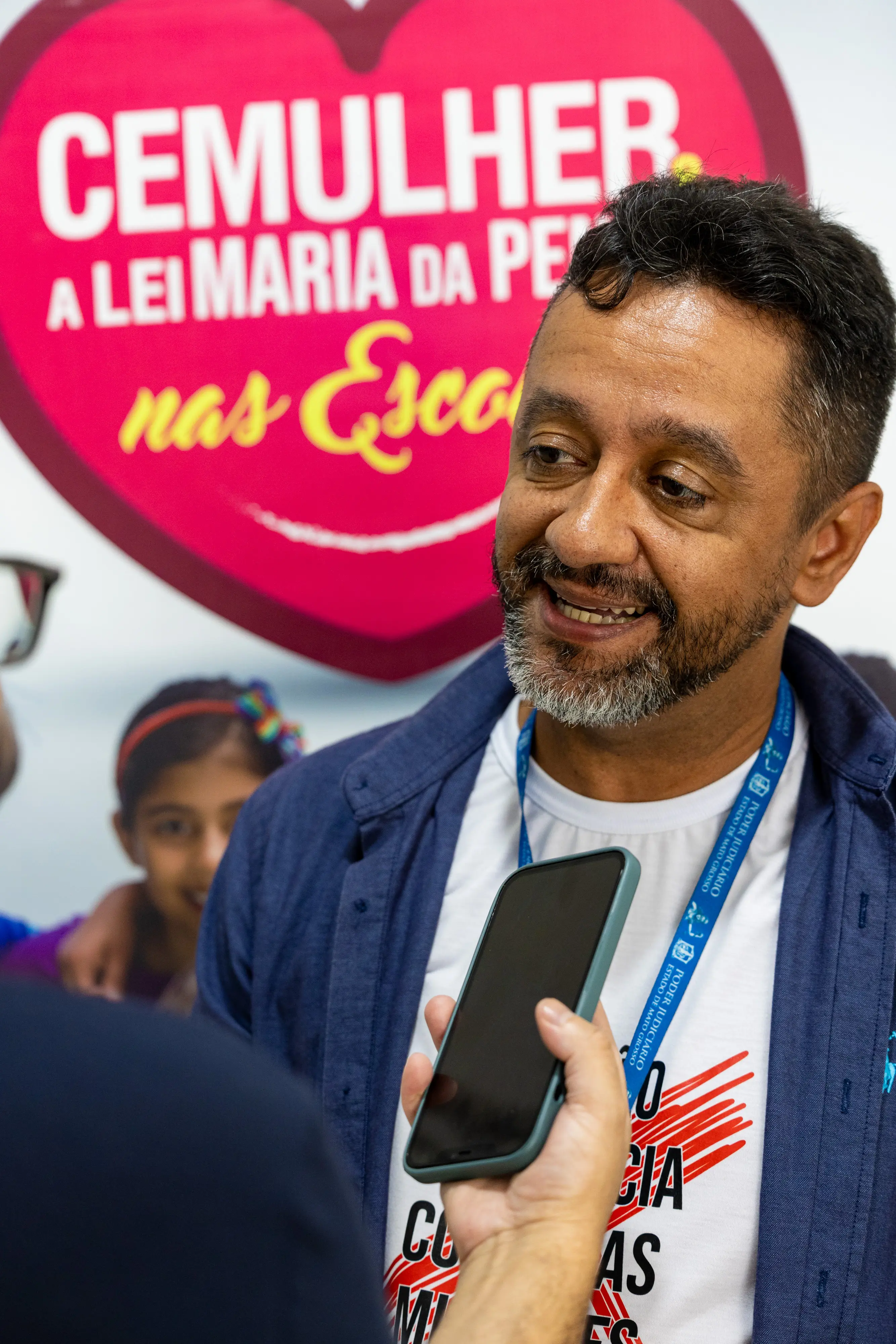 Um homem de barba grisalha e camisa azul sorri enquanto fala ao celular de um entrevistador. Ao fundo, um painel com um coração rosa e os dizeres 