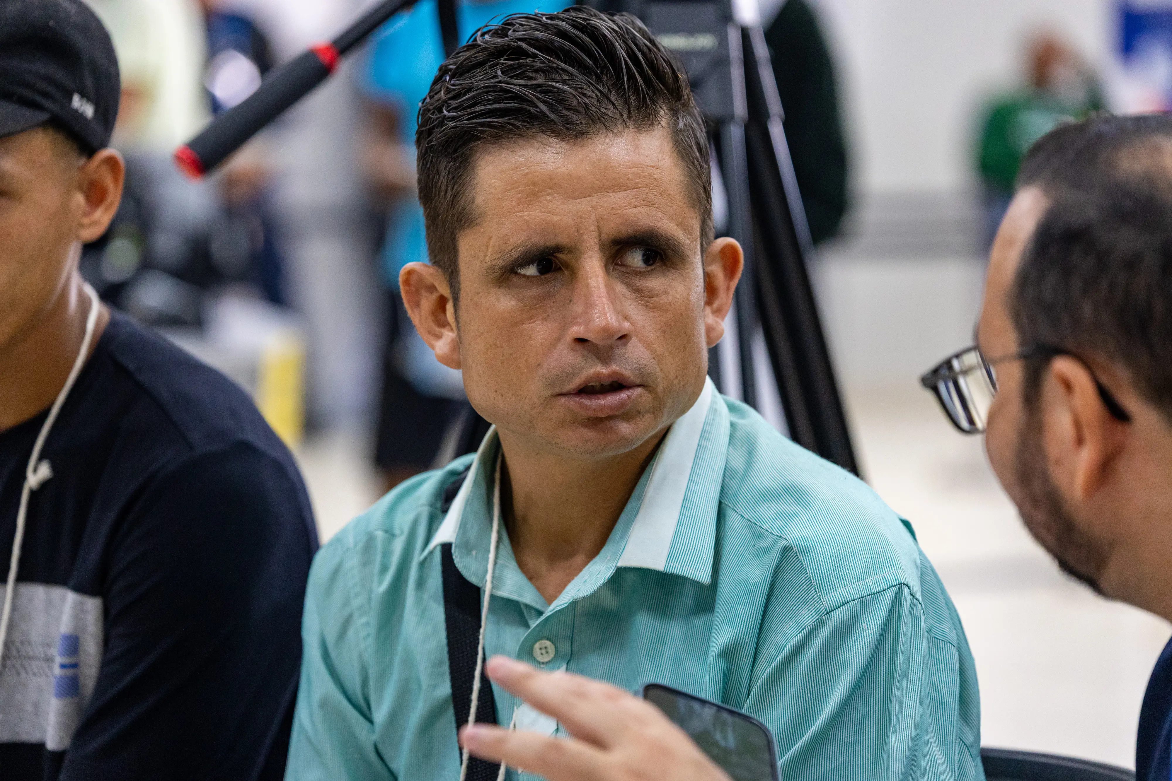 Close de homem de pele clara e cabelos escuros penteados para cima. Ele veste camisa verde clara e olha atentamente para o lado durante uma conversa em um ambiente amplo.