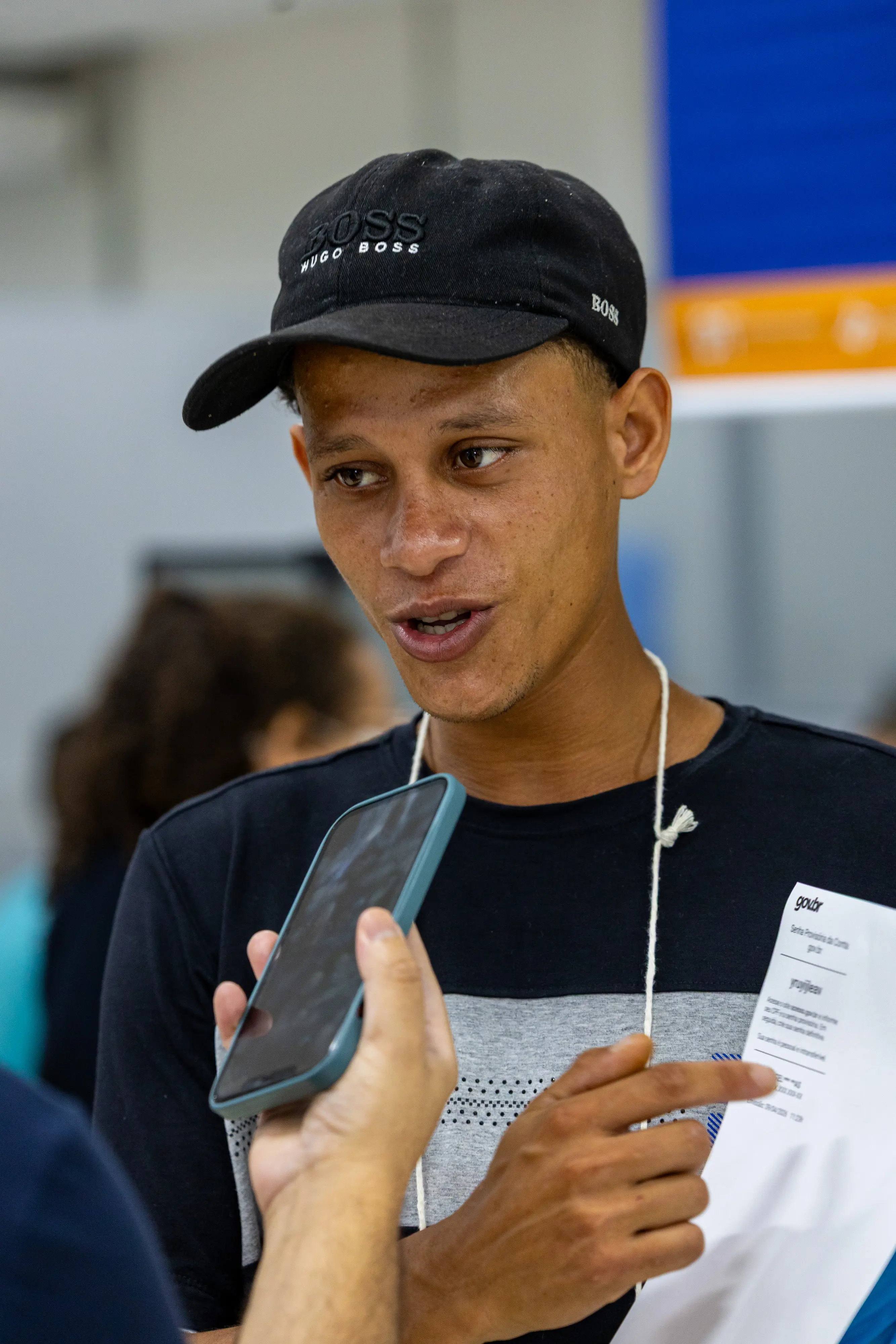 Close de jovem negro de boné preto, falando ao microfone enquanto segura um documento. Ele tem uma expressão concentrada e aponta para o papel durante a entrevista.