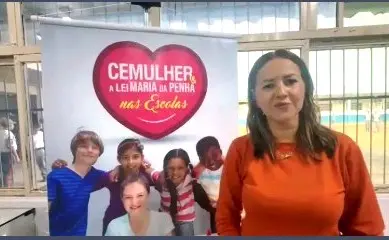 Foto horizontal em plano médio que mostra a professora Fabiana Gomes em pé, ao lado de um banner do projeto Cemulher e a Lei Maria da Penha nas Escolas. ela é uma mulher branca, loira, usando blusa laranjada de manga comprida.