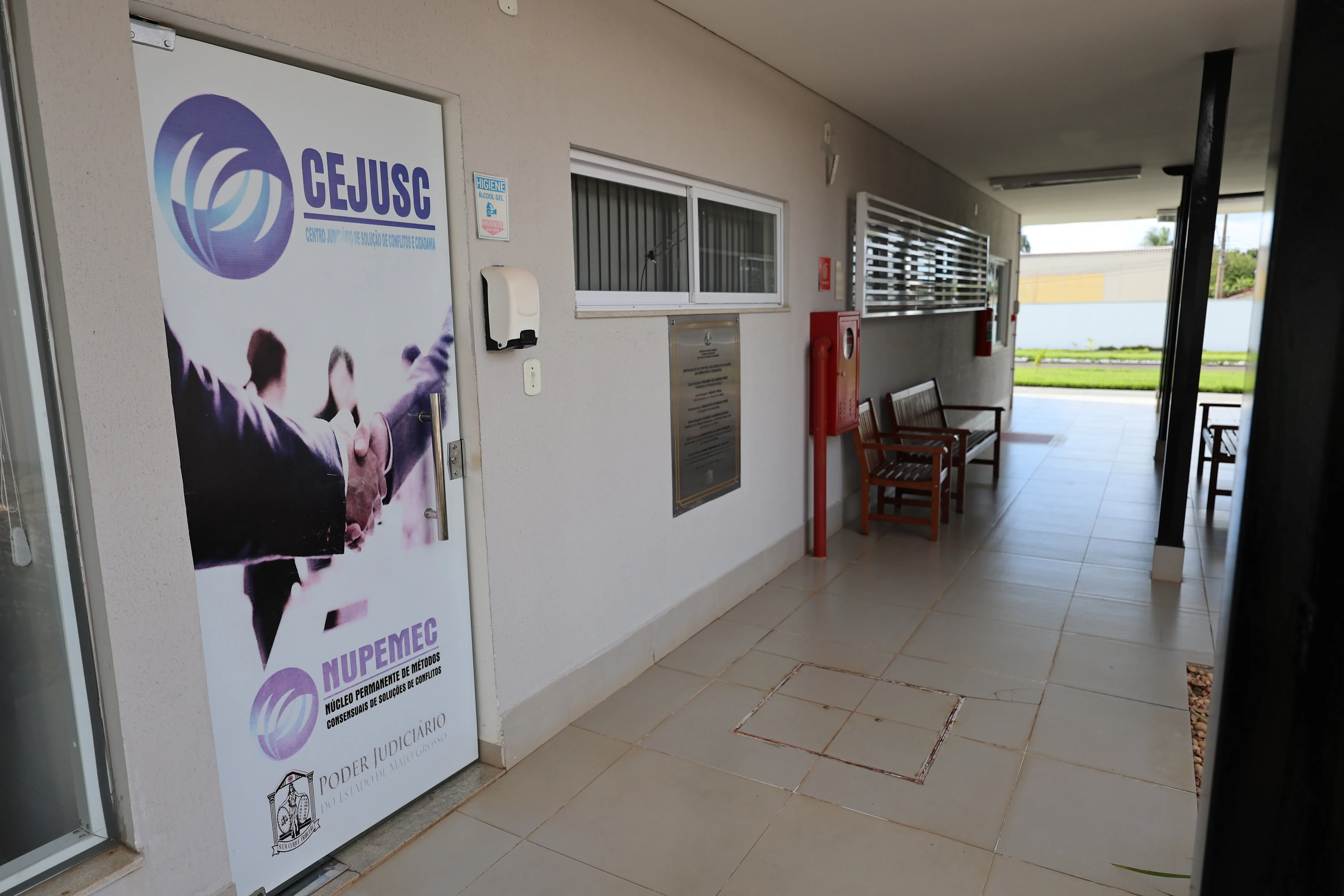 Corredor interno de unidade judiciária com paredes claras e piso cerâmico. À esquerda, painel com identidade visual institucional. Ao fundo, há cadeiras e mesas, com ambiente iluminado e organizado.