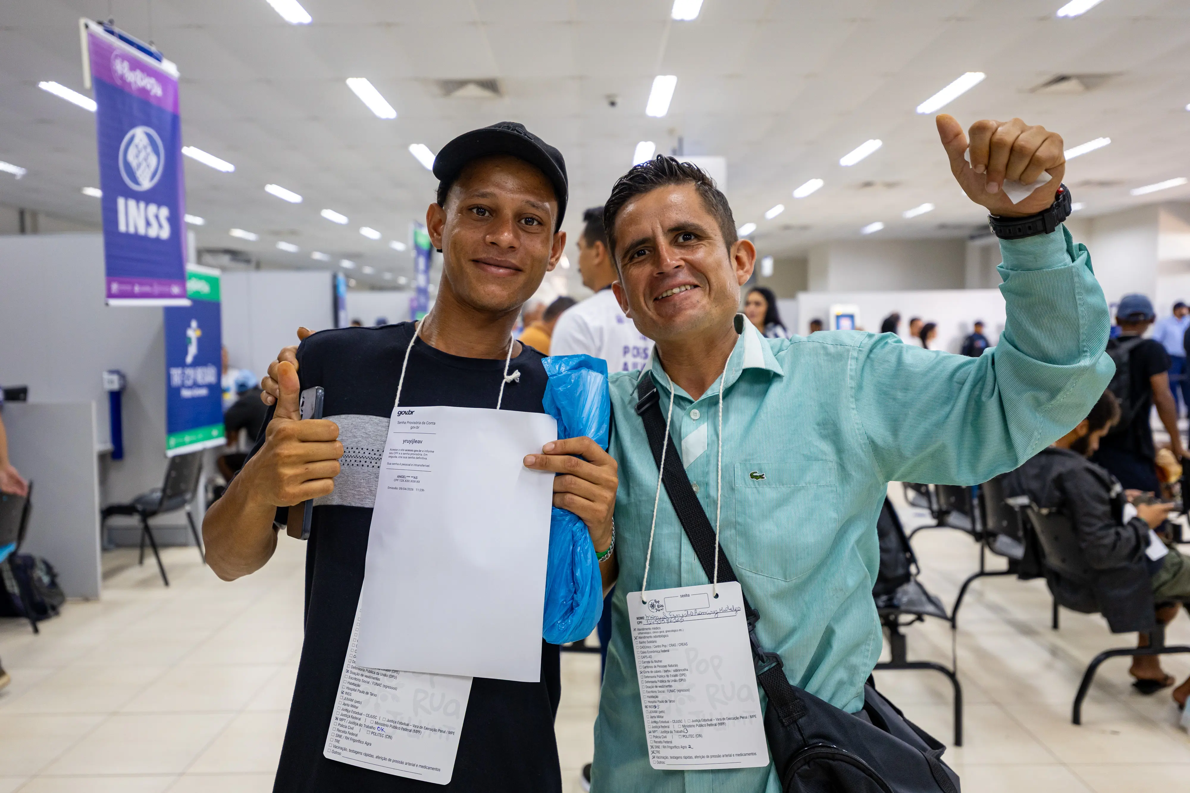Dois homens sorriem abraçados em um mutirão de serviços públicos. O da esquerda, de boné preto, segura um documento oficial; o da direita, de camisa verde, faz sinal de positivo.