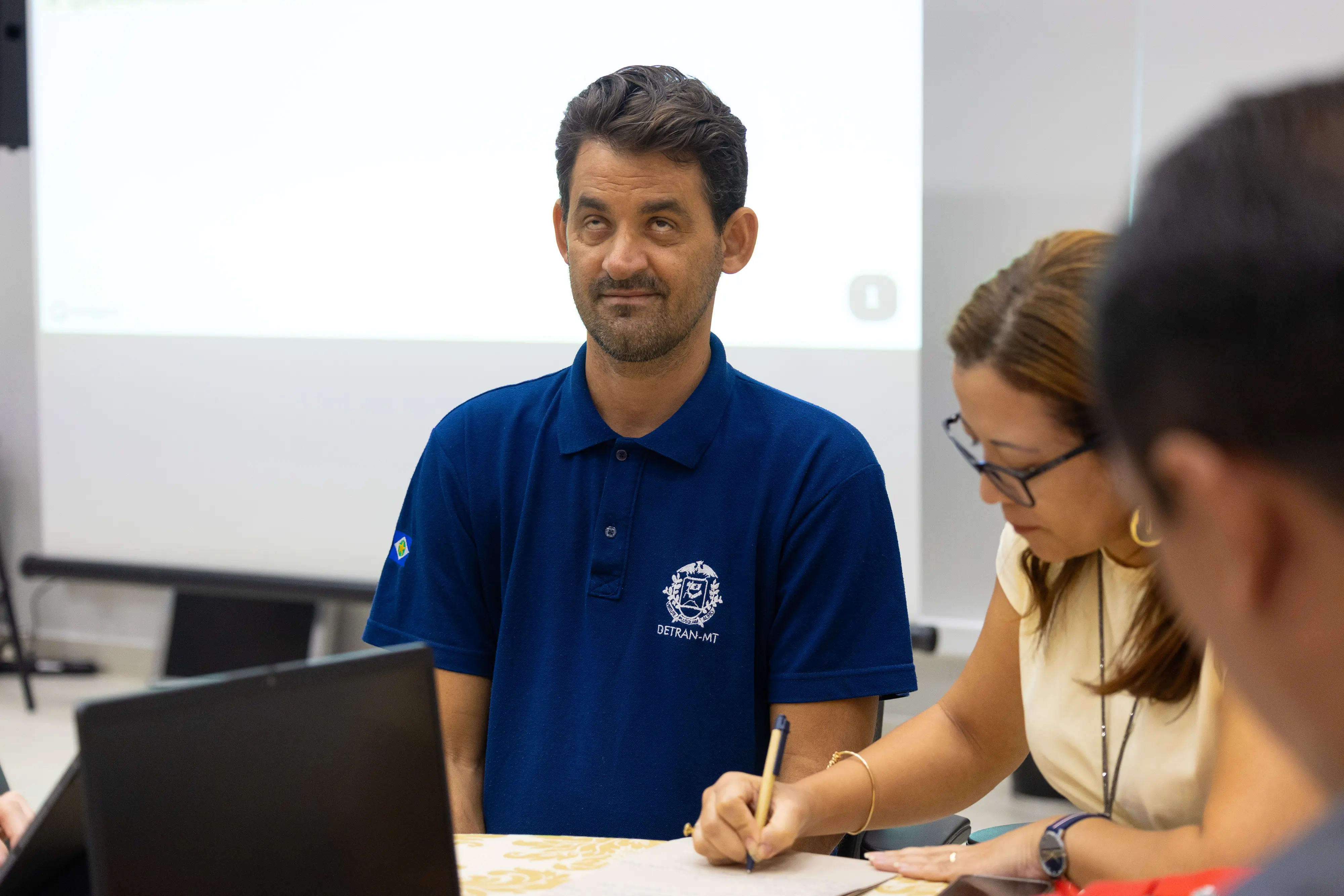Homem de camisa azul fala sorrindo durante oficina participativa. Pessoas ao redor acompanham a conversa.