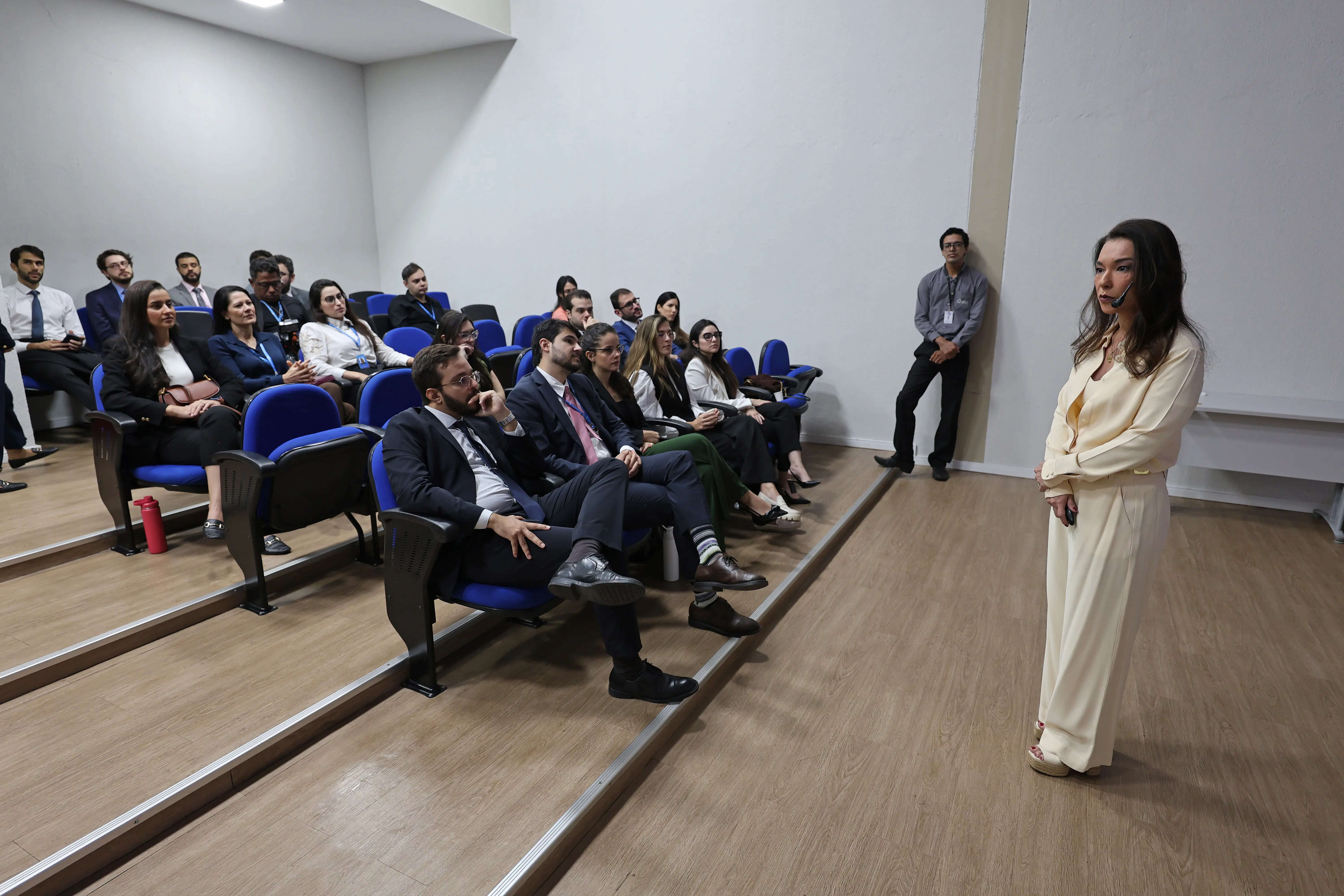 Foto horizontal em plano aberto que mostra novos magistrados sentados em um auditório, ouvindo palestra da juíza Hanae Yamamura, que está em pé à frente.