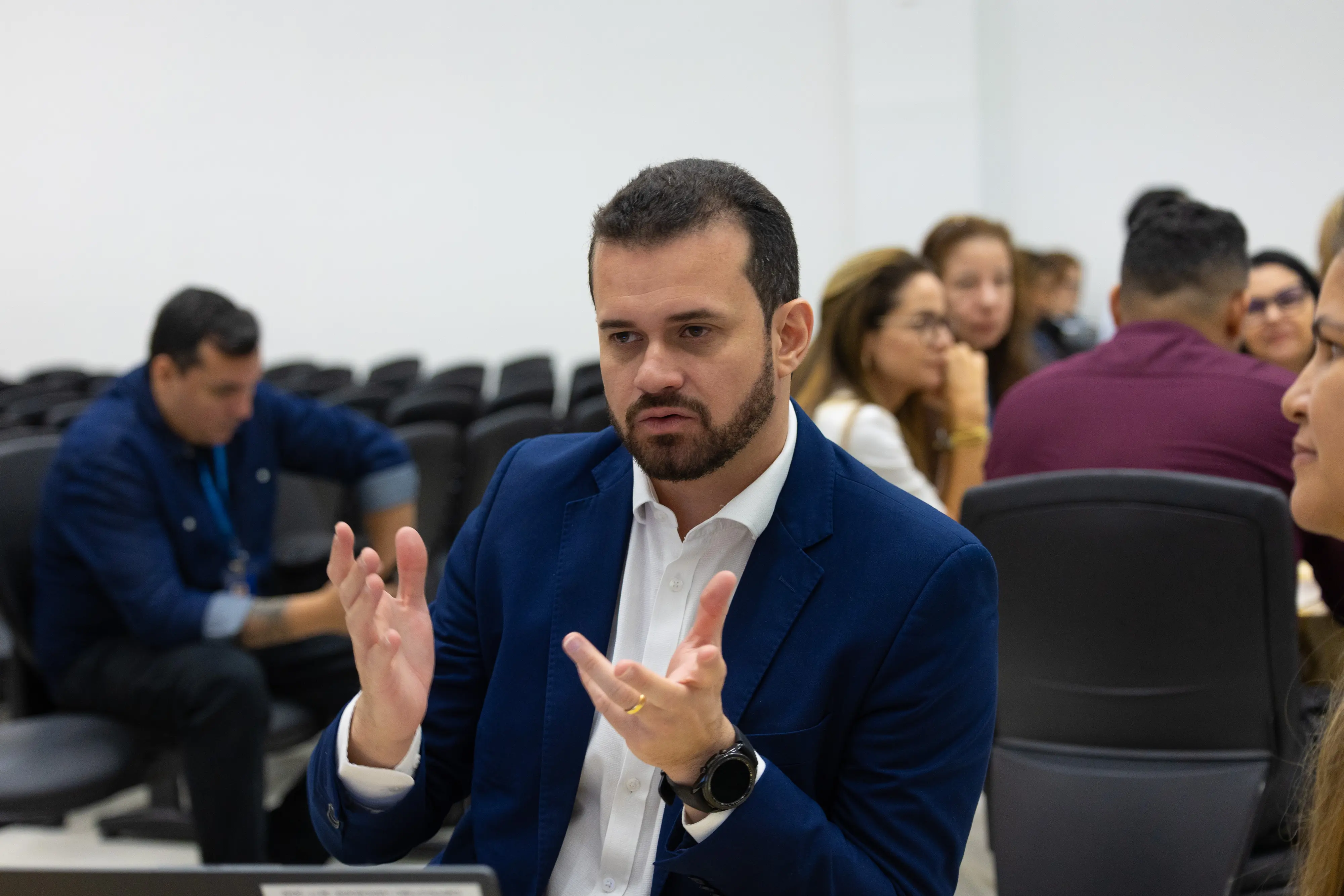 Homem de blazer azul participa de roda de conversa durante oficina. Participantes acompanham atentos ao fundo.