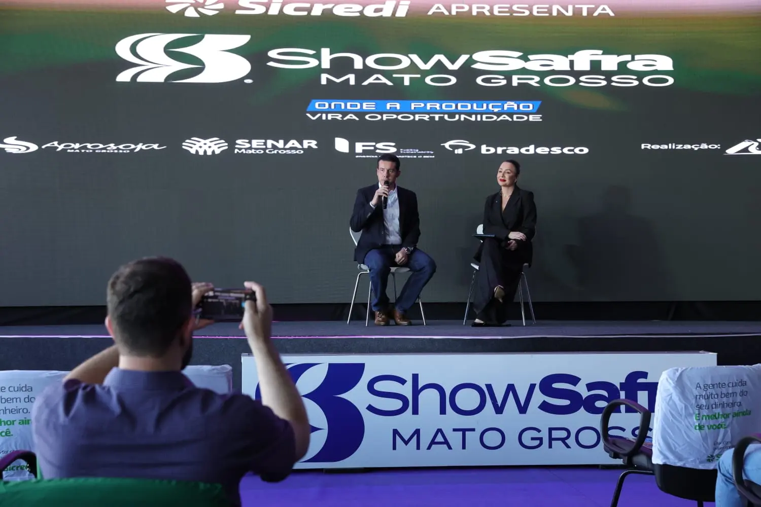 Homem e mulher palestram em palco sob telão do 