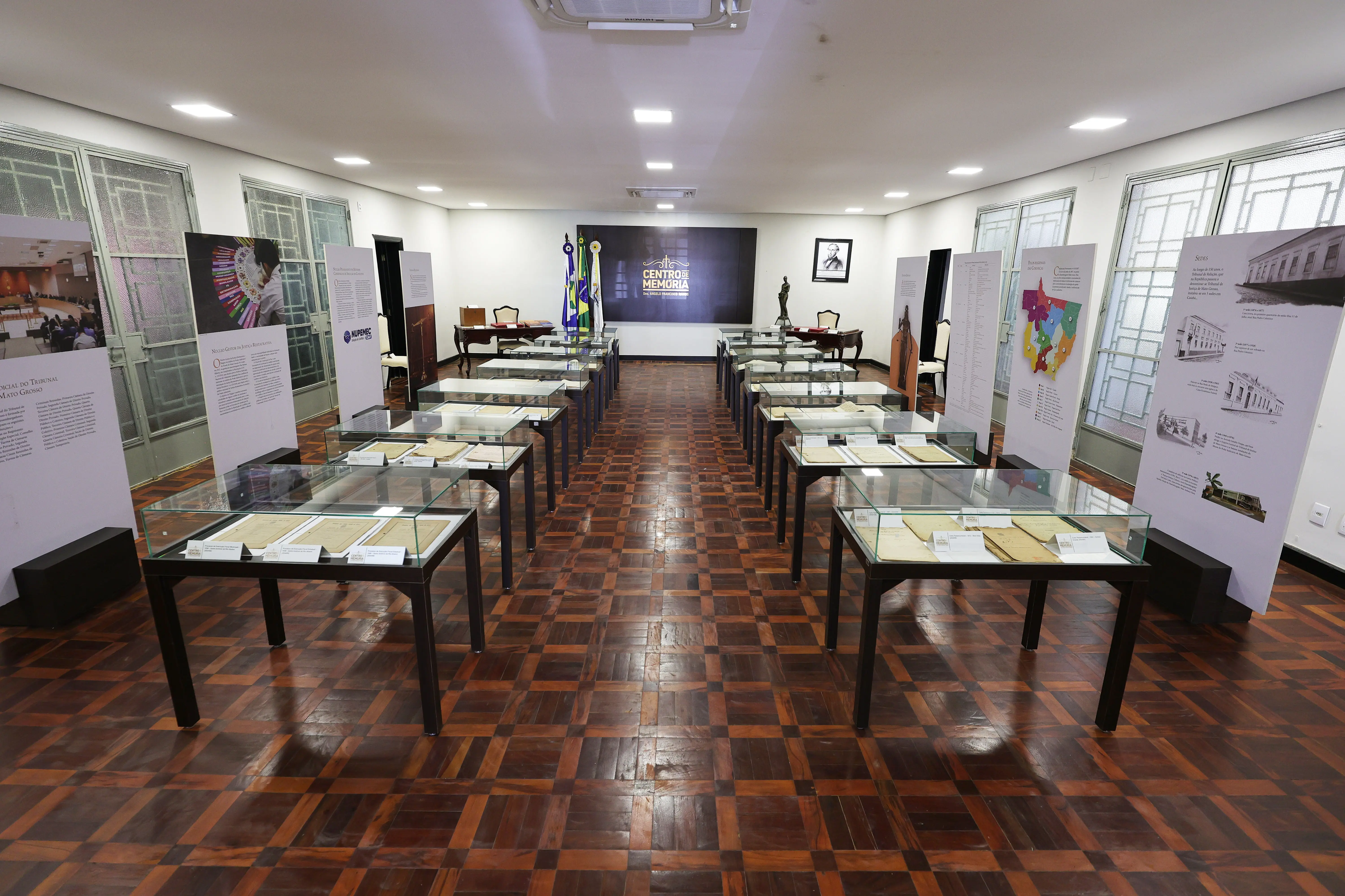 Visão ampla de uma exposição histórica em sala com piso de madeira em padrão xadrez. Várias vitrines de vidro retangulares estão alinhadas sobre mesas escuras. Nas laterais, painéis informativos altos. Ao fundo, um painel escuro com o logo 'CENTRO DE MEMÓRIA', bandeiras e um busto.