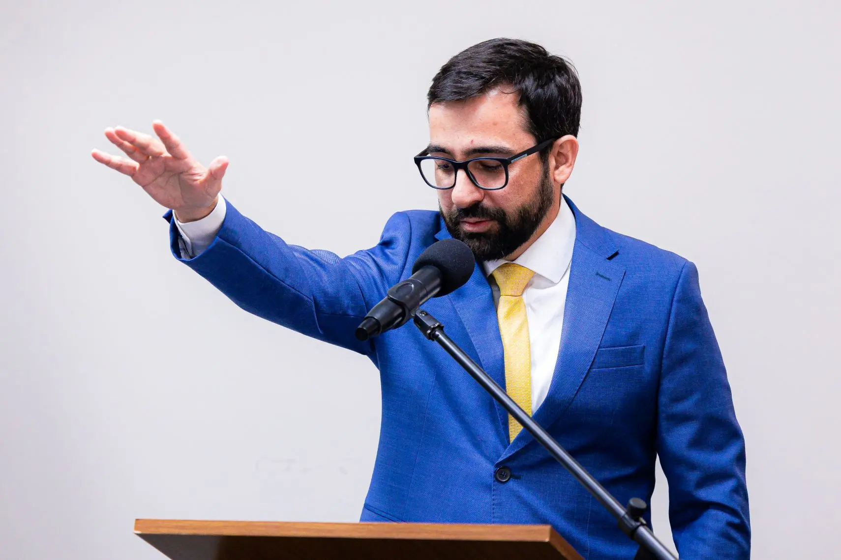 Foto horizontal que mostra o juiz Eduardo Henrique Minosso fazendo juramento em sua posse como juiz do TJMT. Ele é um homem branco de cabelos e barba escuros, usando terno azul, gravata amarela, óculos de grau. Ele fala ao microfone e está com o braço direito estendido.