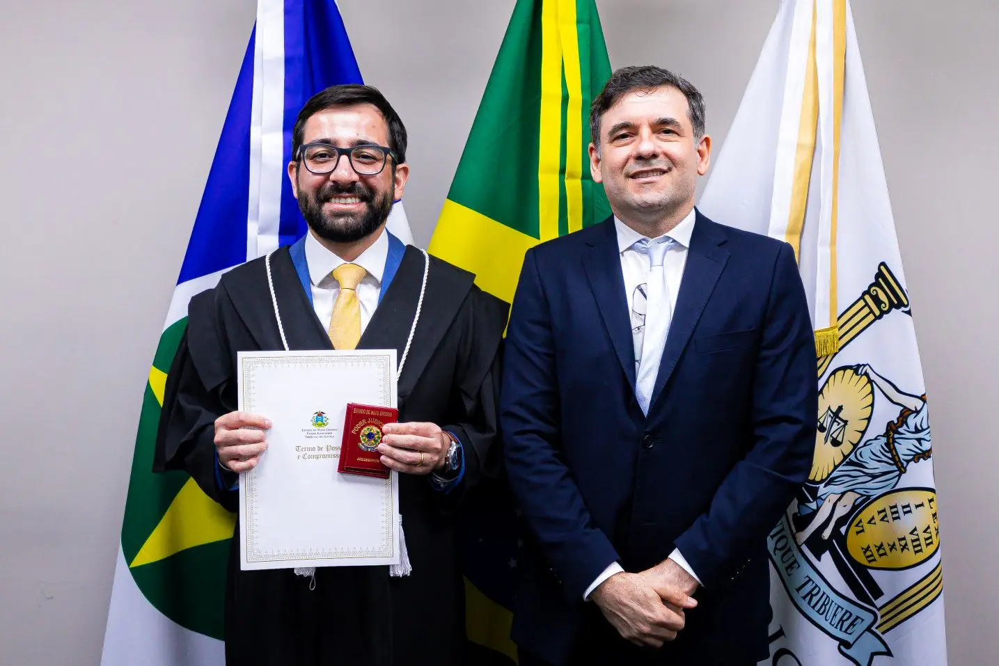 Foto horizontal que mostra o juiz Eduardo Henrique Minosso posando para foto segurando seu termo de posse e carteira funcional do TJMT, togado, ao lado de outro homem que usa terno e grata. Atrás deles aparecem as bandeiras do Brasil, de Mato Grosso e do Judiciário de Mato Grosso.