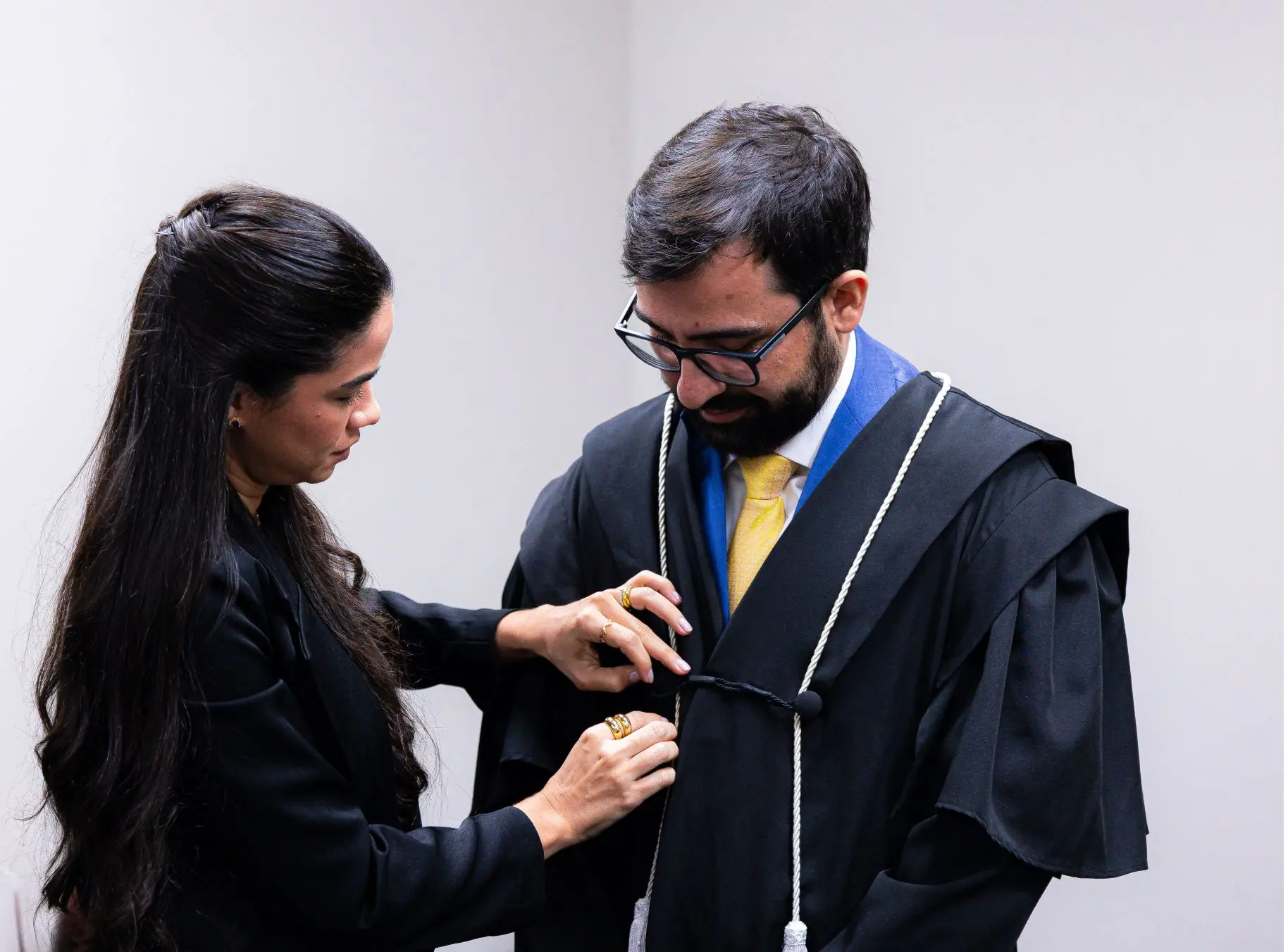 Foto horizontal que mostra o juiz Eduardo Henrique Minosso em pé, com sua esposa amarrando o cordão de sua toga de juiz. Ambos usam preto. Ambos tem cabelos castanhos escuros. Ela tem cabelos muito longos e ele usa óculos de grau.