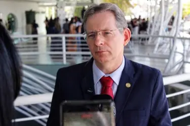 Pessoa de terno escuro e gravata clara conversa com repórter, com microfone e celular visíveis em primeiro plano. Ao fundo, estrutura com corrimão metálico e pessoas desfocadas.