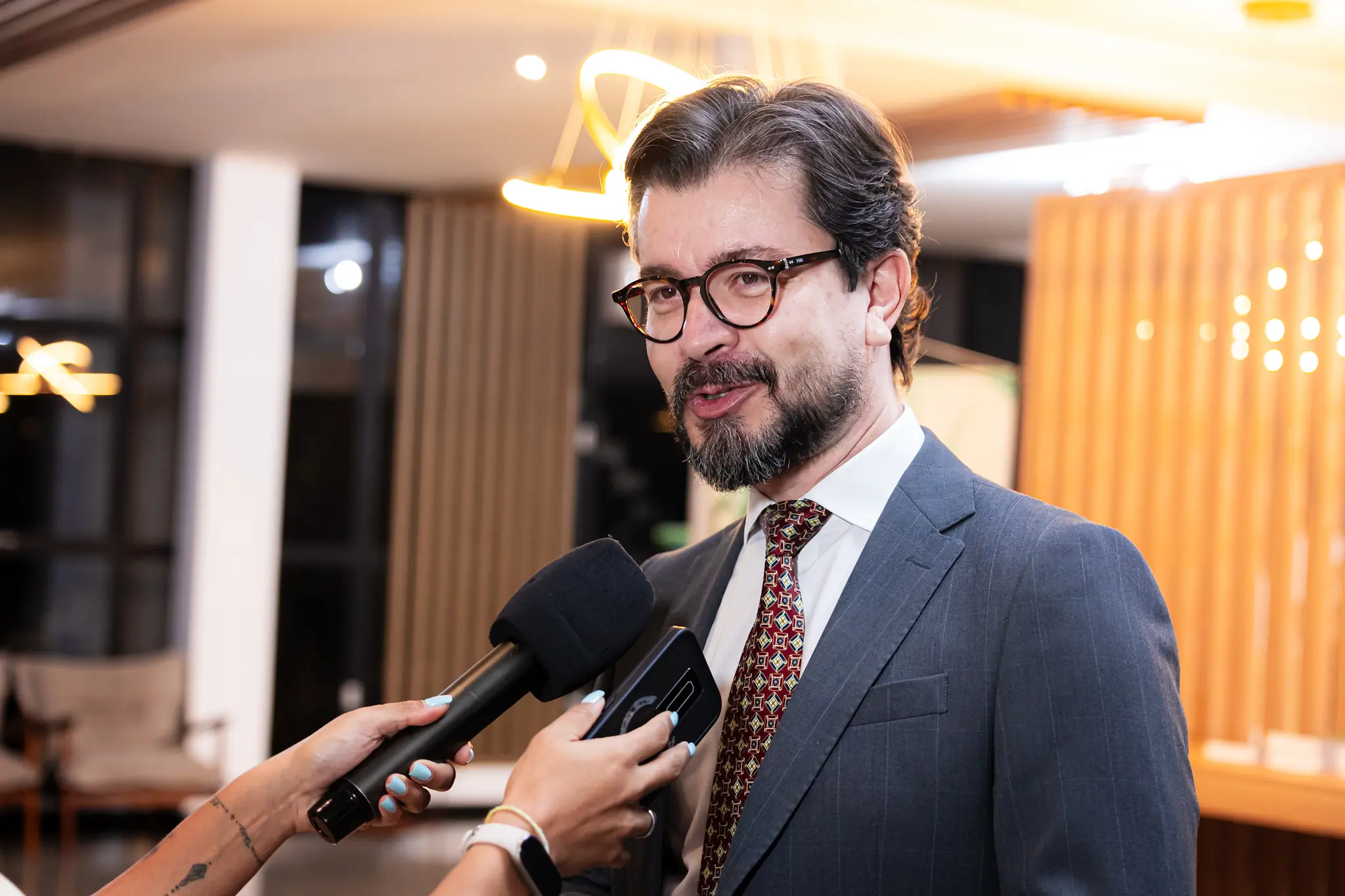 Homem de óculos, barba e cabelos castanhos com grisalho concede entrevista. Ele usa terno cinza, camisa branca e gravata estampada em tons vermelhos.