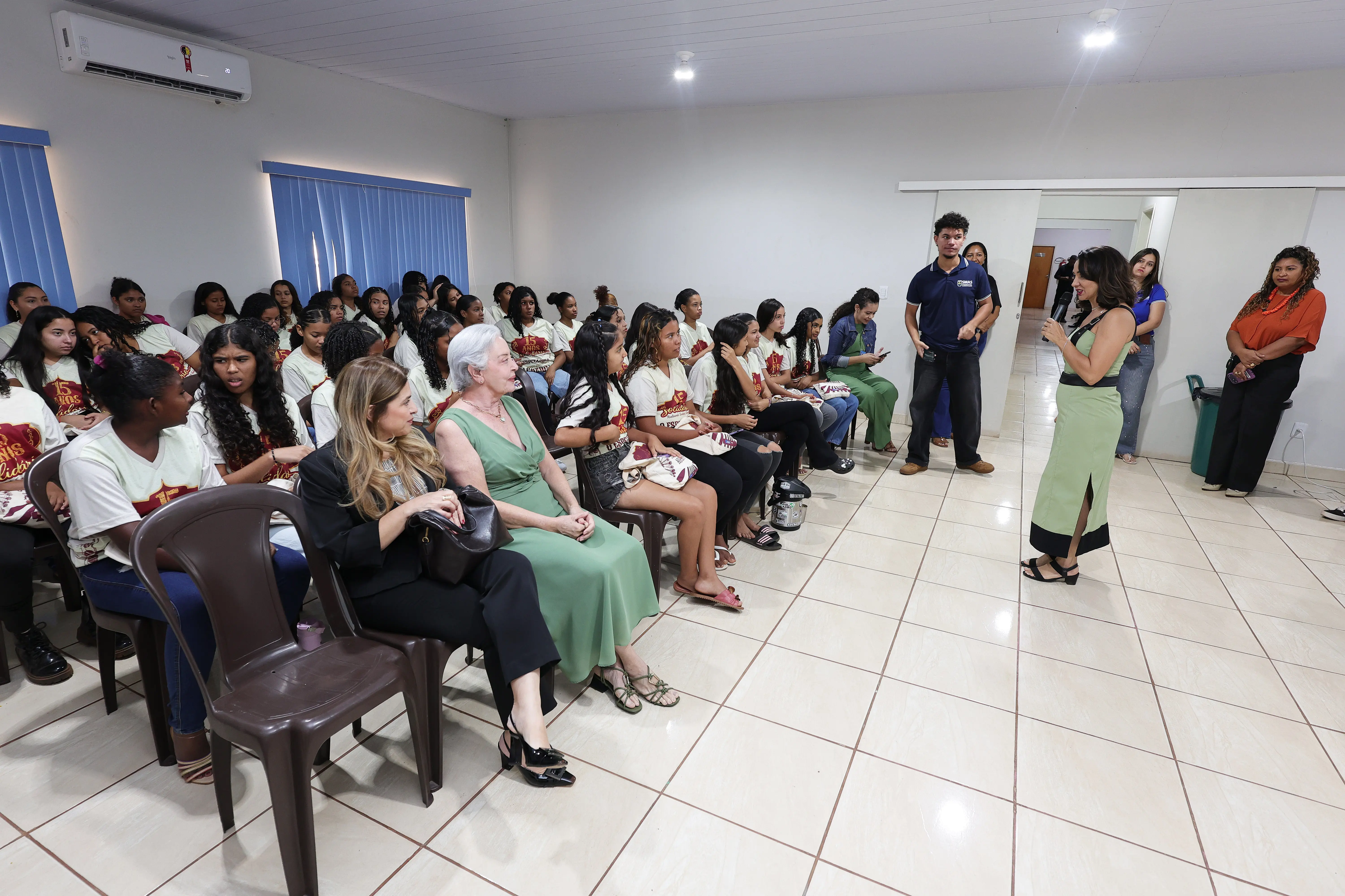 Mulher de vestido verde fala ao microfone em pé diante de uma plateia de jovens e convidadas sentadas. Outras pessoas acompanham a palestra em pé ao fundo.