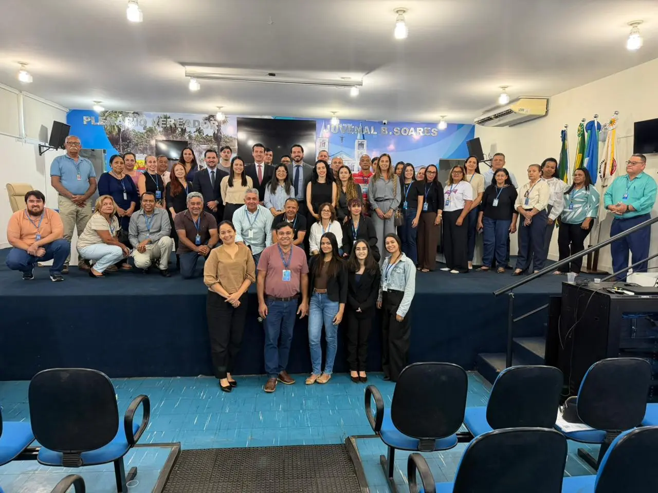 Foto em grupo com cerca de quarenta pessoas reunidas no plenário da Câmara Municipal. A maioria está de pé sobre o palco e algumas poucas estão posicionadas no chão, à frente. Ao fundo, painel azul com a inscrição 