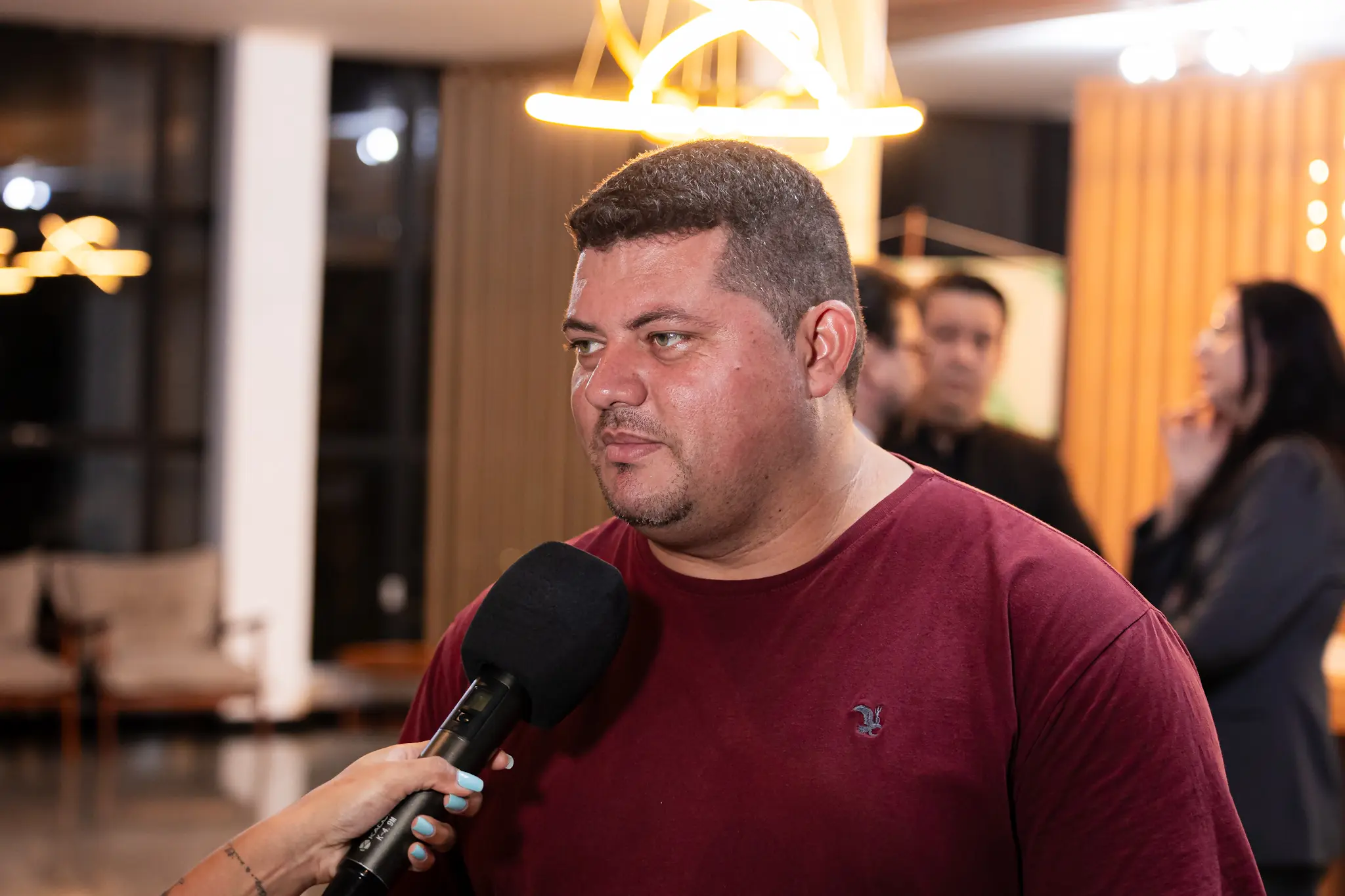 Homem de cabelo curto e barba rala, veste camisa bordô e olha para a esquerda, pensativo. Ele segura um microfone preto, sendo entrevistado em um ambiente de evento com iluminação quente.