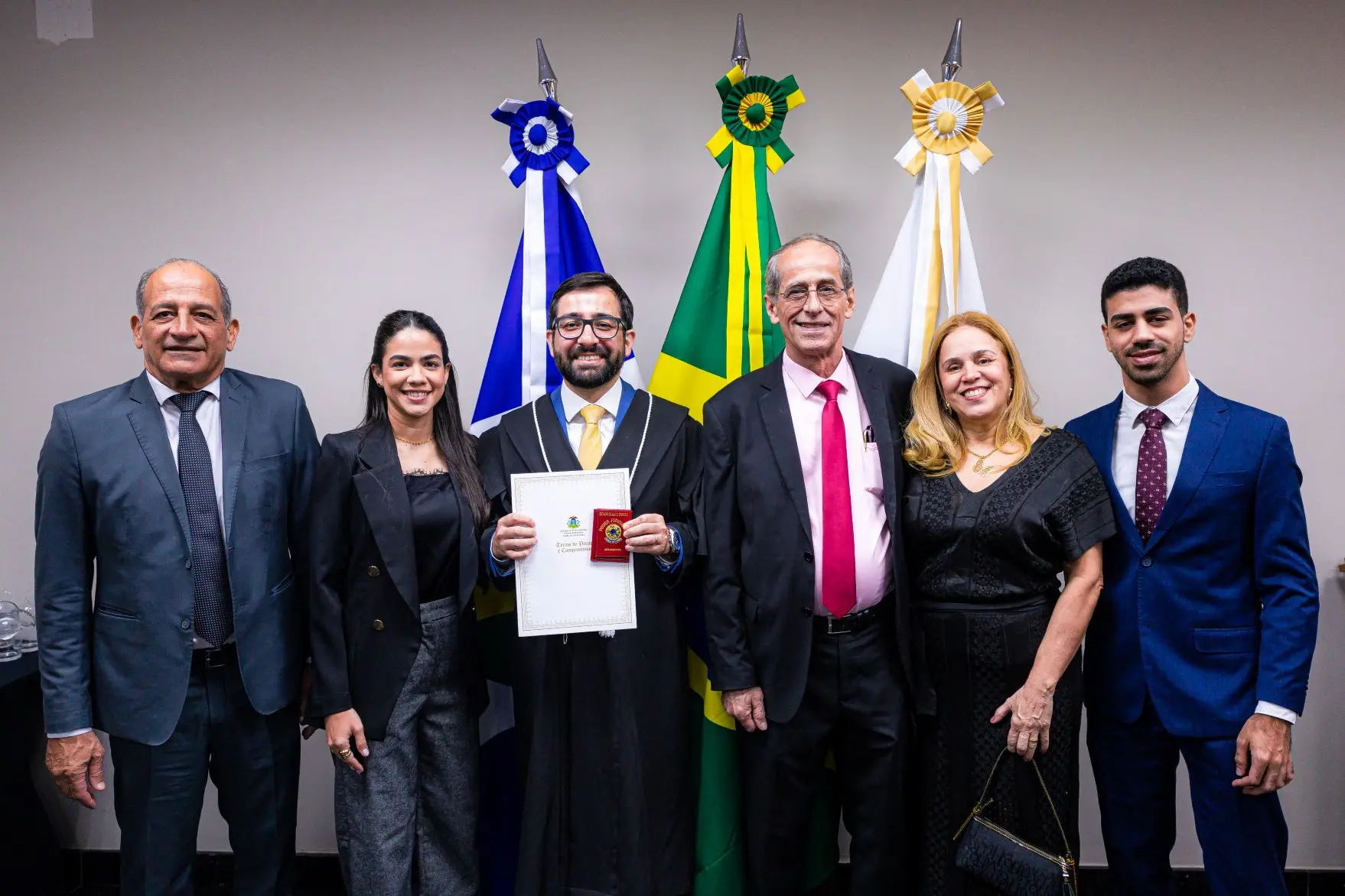 Foto horizontal que mostra o juiz Eduardo Henrique Minosso, usando toga e mostrando sua carteira funcional e termo de posse no TJMT, ao lado de sua esposa, pais e demais familiares.