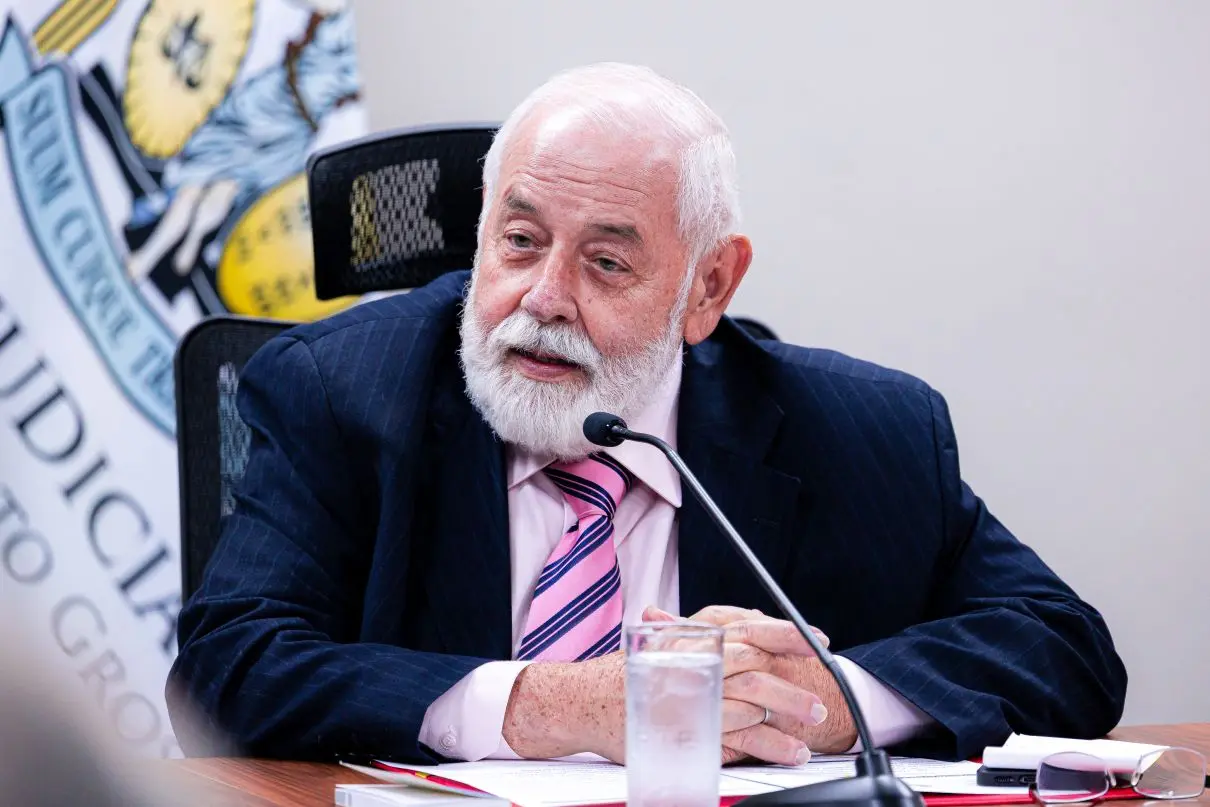 Foto horizontal que mostra o presidente do TJMT, desembargador José Zuquim Nogueira, sentado, com os braços sobre a mesa e falando ao microfone. Ele é um senhor branco, de olhos azuis, cabelo e barba brancos, usando camisa rosa clara, gravara rosa com listras azuis, terno azul marinho risca-de-giz.