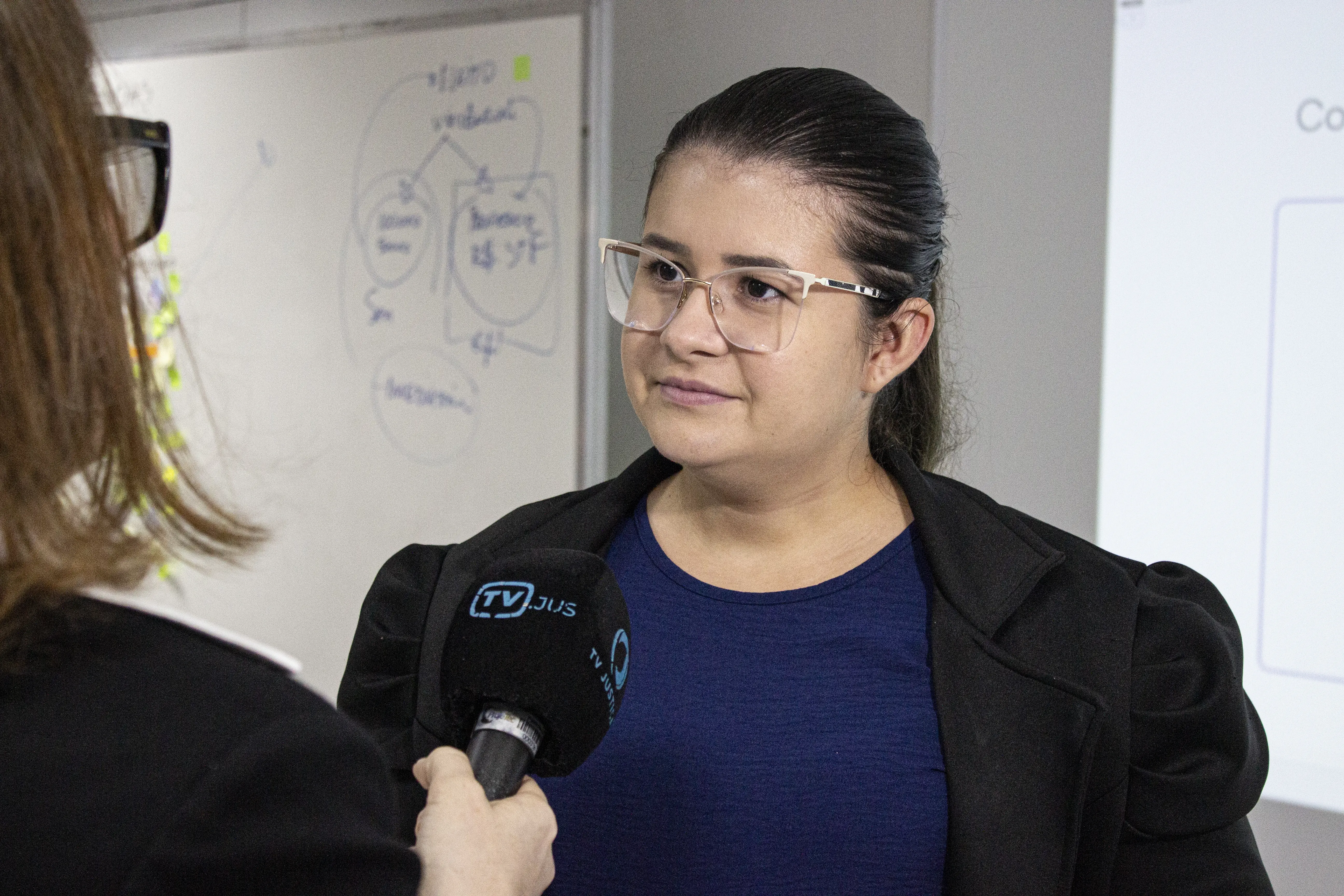 Mulher de óculos e cabelo preso participa de entrevista em ambiente interno. À frente dela, uma pessoa segura microfone; ao fundo há quadro branco com anotações.