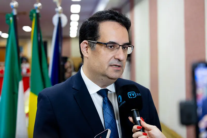 Foto horizontal colorida, em plano médio, que mostra o juiz Tulio Duailibi, durante entrevista à TV Justiça, no auditório do Ministério Público. Ele é um homem branco, alto, magro, de olhos e cabelos pretos, usando camisa branca, terno e gravata azul marinho e óculos de grau.