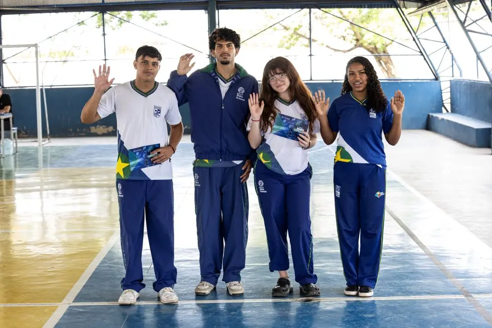 Foto horizontal colorida em plano aberto que mostra os estudantes Felipe, Murilo, Ana Vitória e Daviela em pé, sorrindo e acenando para a foto, na quadra da escola. Todos usam uniforma da escola (calça e camiseta ou moletom). 