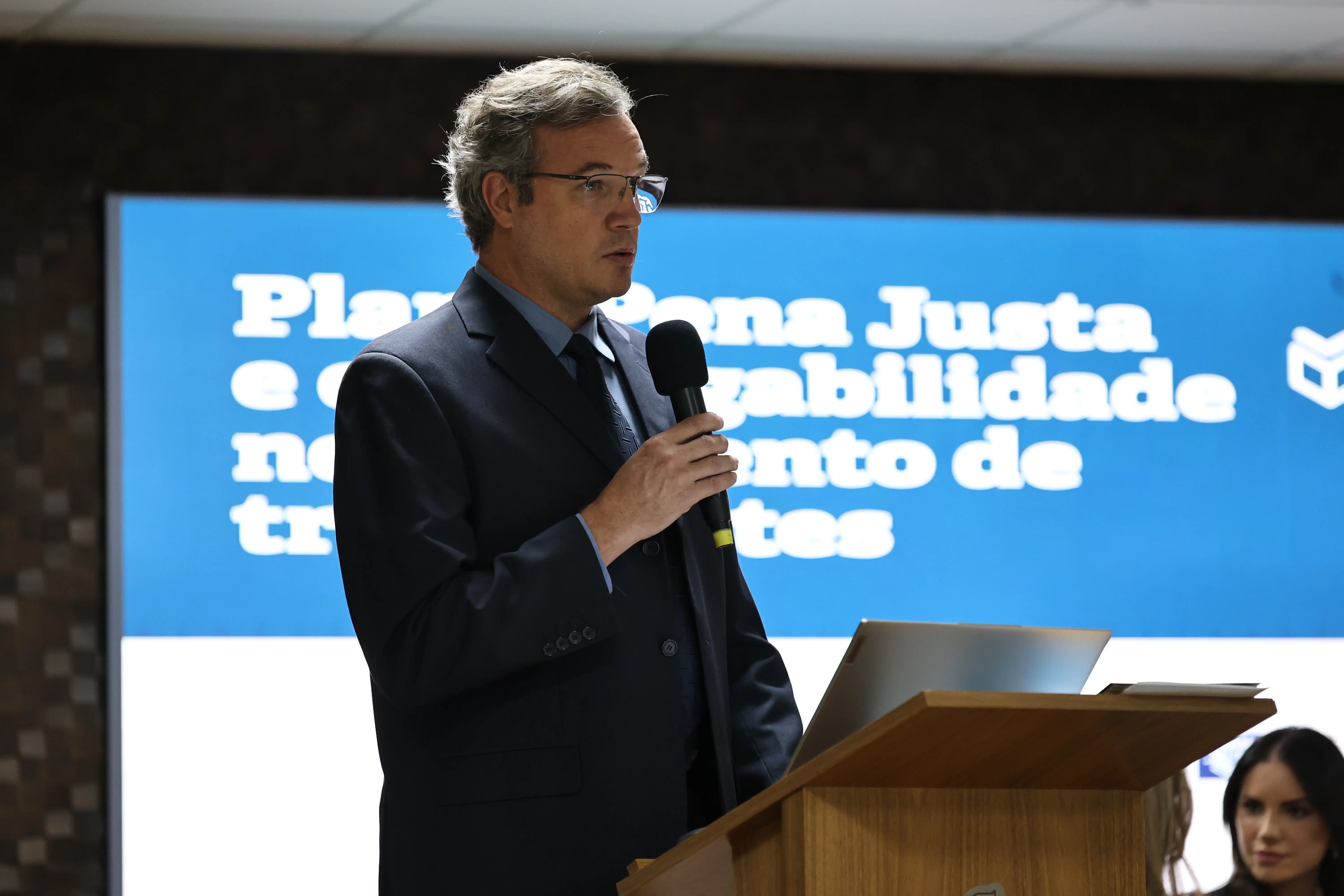 Homem branco, com cabelos grisalhos. Ele usa óculos, terno preto, camisa azul e gravata escura. Ele segura um micronfe e fala em frente a um telaão azul com letras brancas.