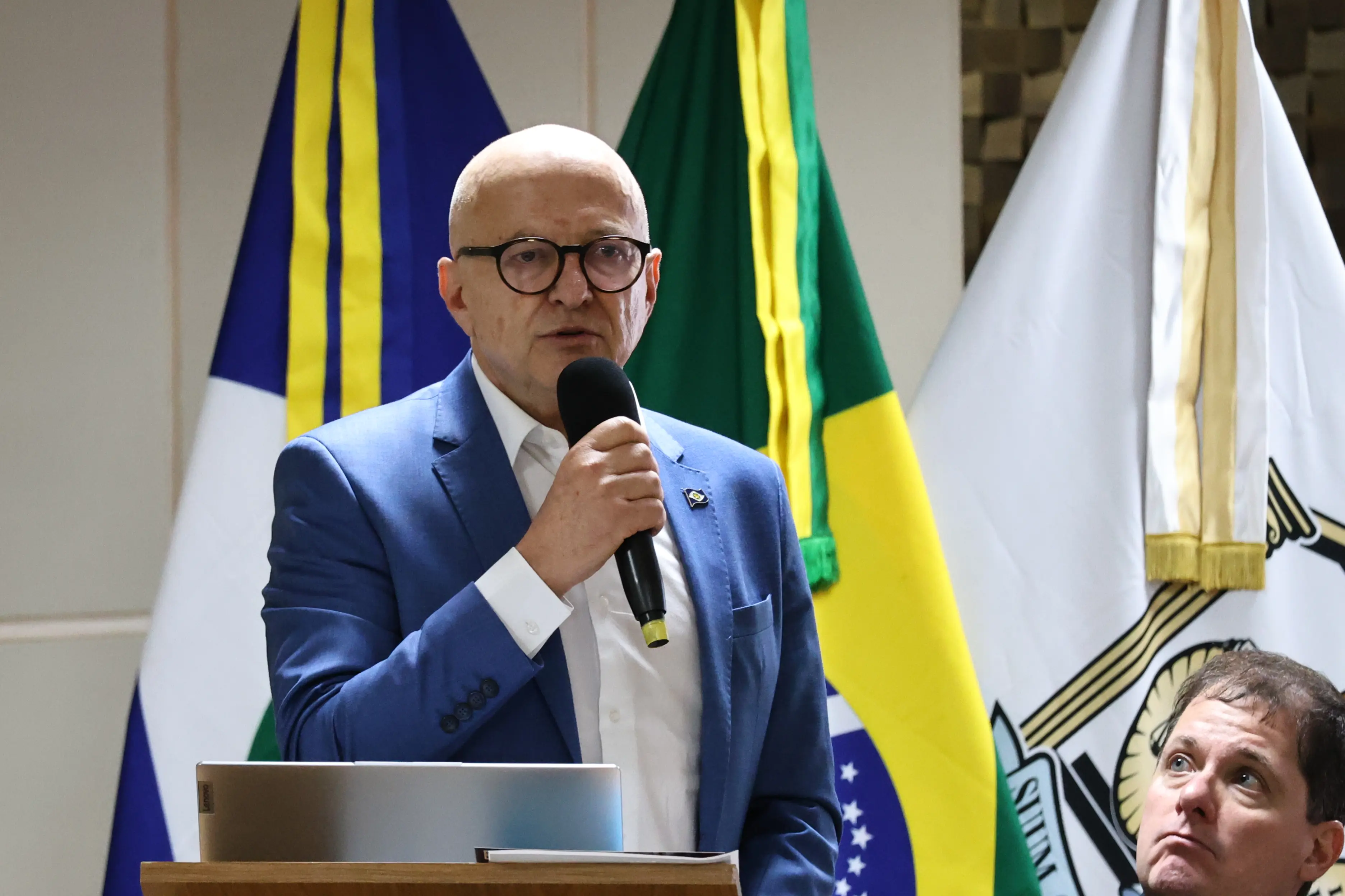 Homem branco, careca. Ele usa óculos, ternno azul e camisa branca. Fala ao microfone, atrás de um púlpito de madeira, em frente a bandeiras do Brasil, Mato Grosso e do Poder Judiciário.