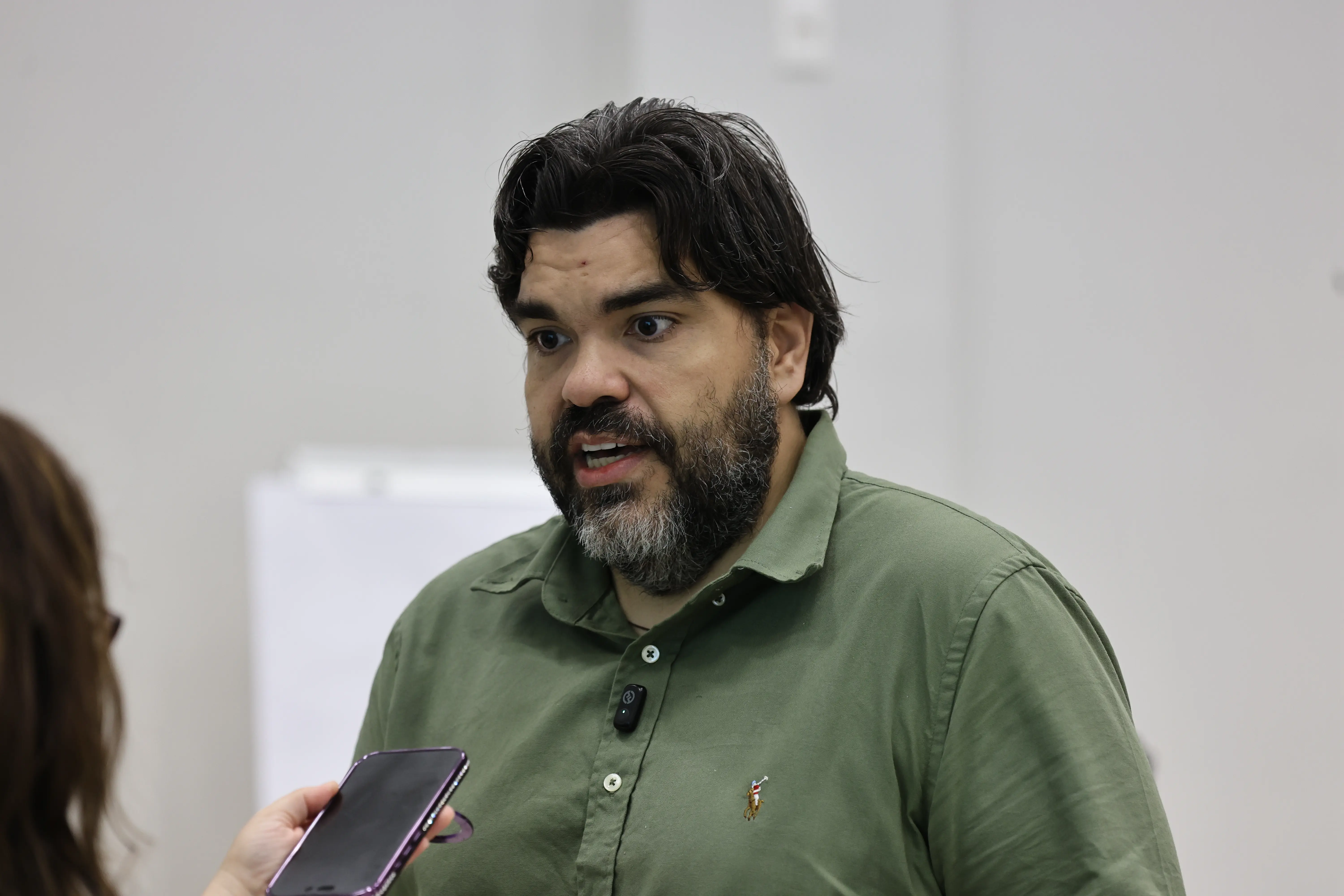 Homem concede entrevista, falando a um celular usado como gravador. Expressão séria indica posicionamento. Ambiente interno neutro reforça contexto institucional.