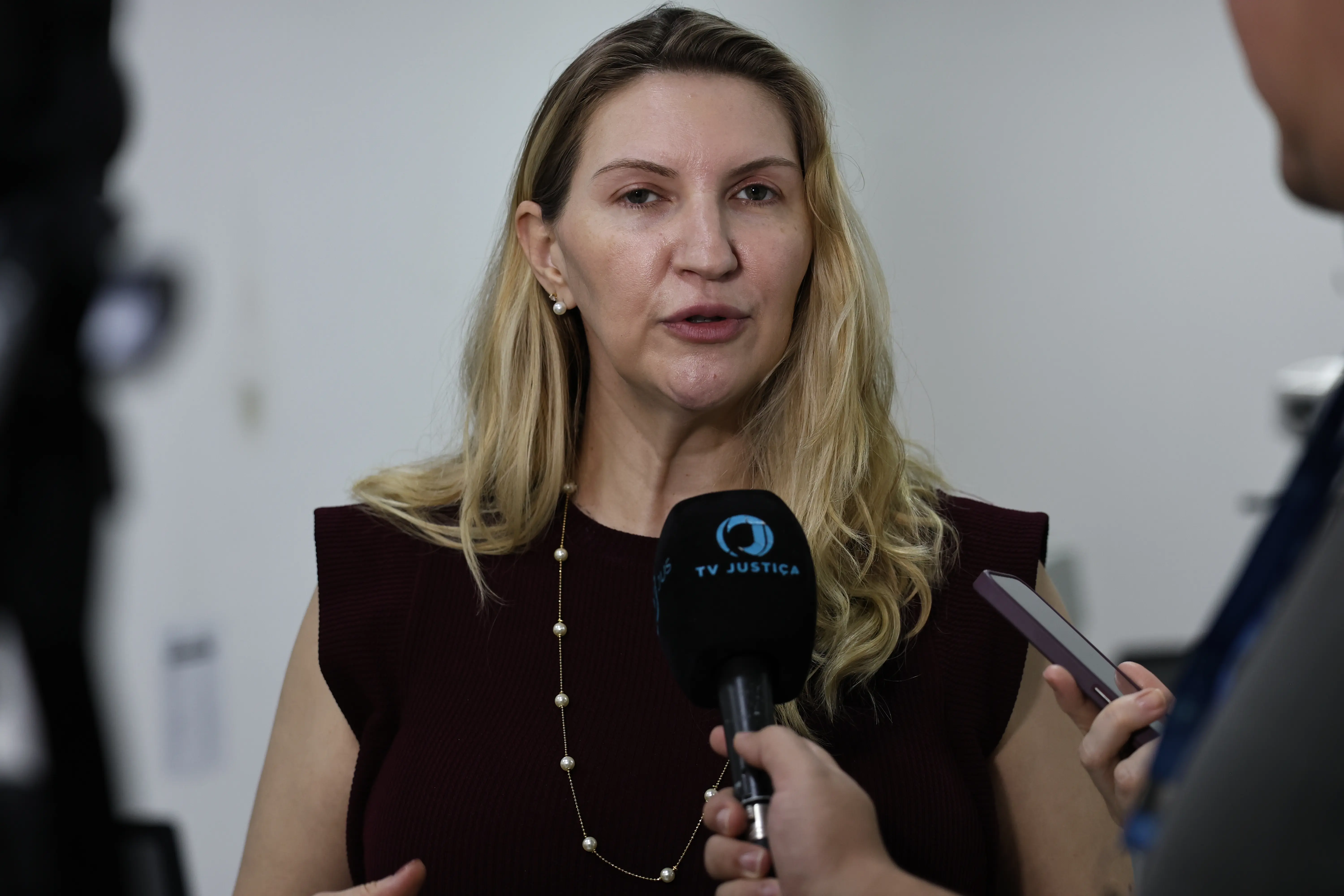 Mulher loira de blusa escura e colar de pérolas fala ao microfone da "TV JUSTIÇA". Ela gesticula com as mãos enquanto é entrevistada em um ambiente interno de paredes claras.