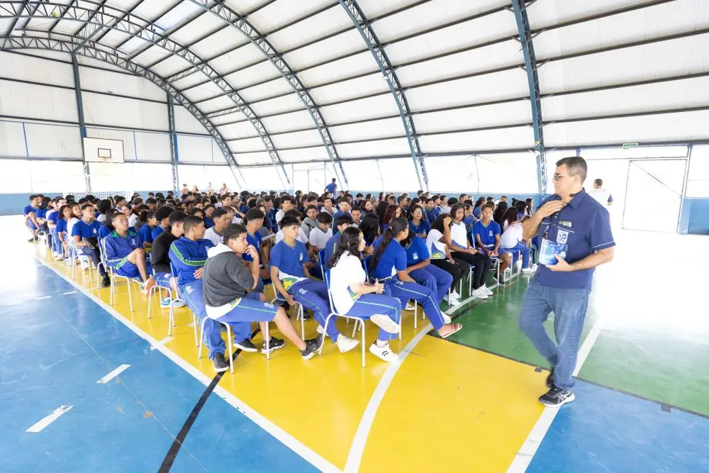 Foto horizontal colorida em plano aberto que mostra mais de 200 alunos uniformizados sentados em cadeiras na quadra da Escola Estadual Leovegildo de Melo.