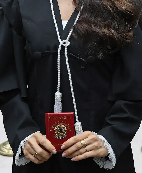 Foto vertical em plano fechado que mostra detalhe da toga de uma juíza, que está em pé, segurando sua carteira funcional. 