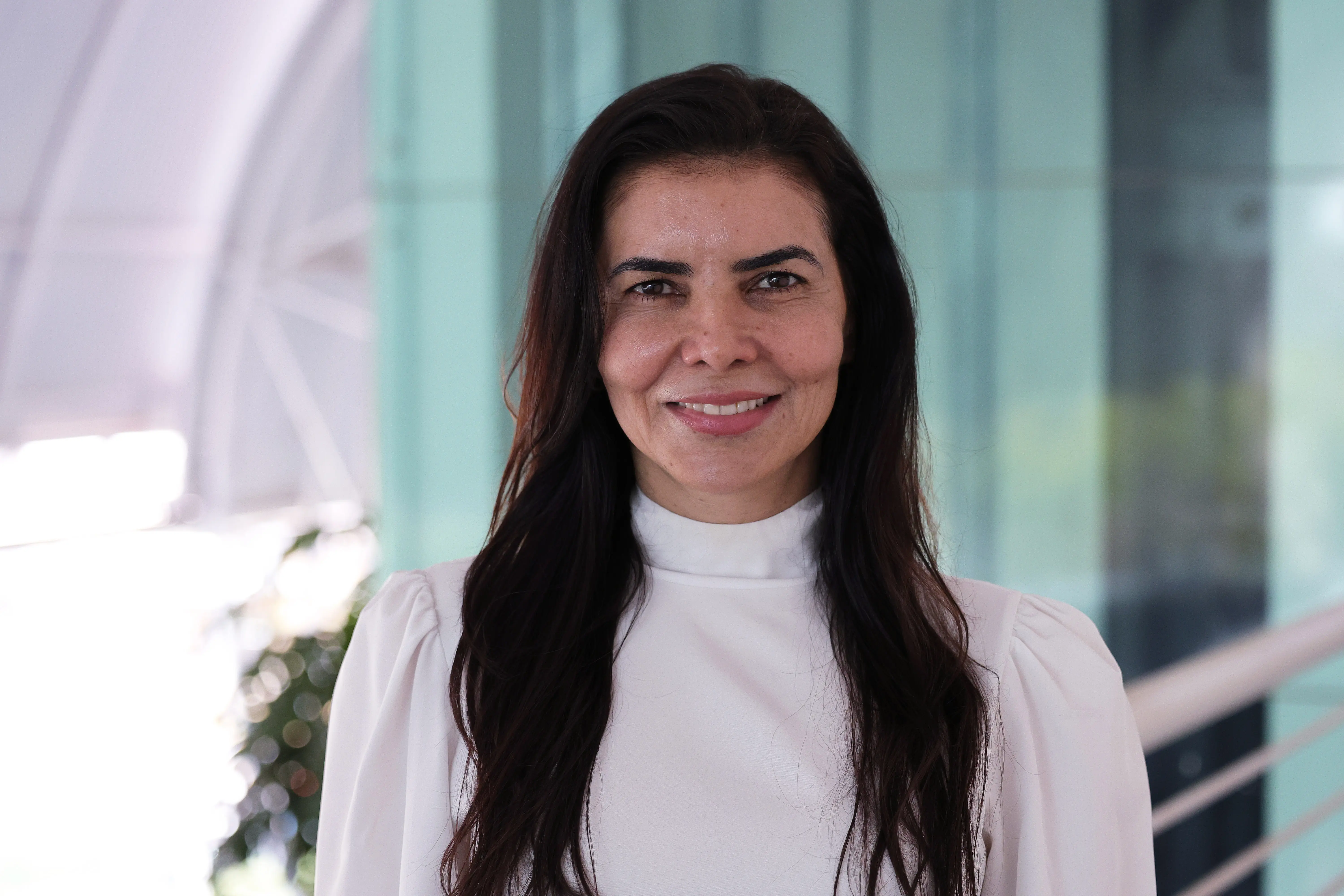 Retrato de uma mulher de cabelos longos escuros e blusa branca de gola alta, sorrindo para a câmera em um corredor com paredes de vidro ao fundo.